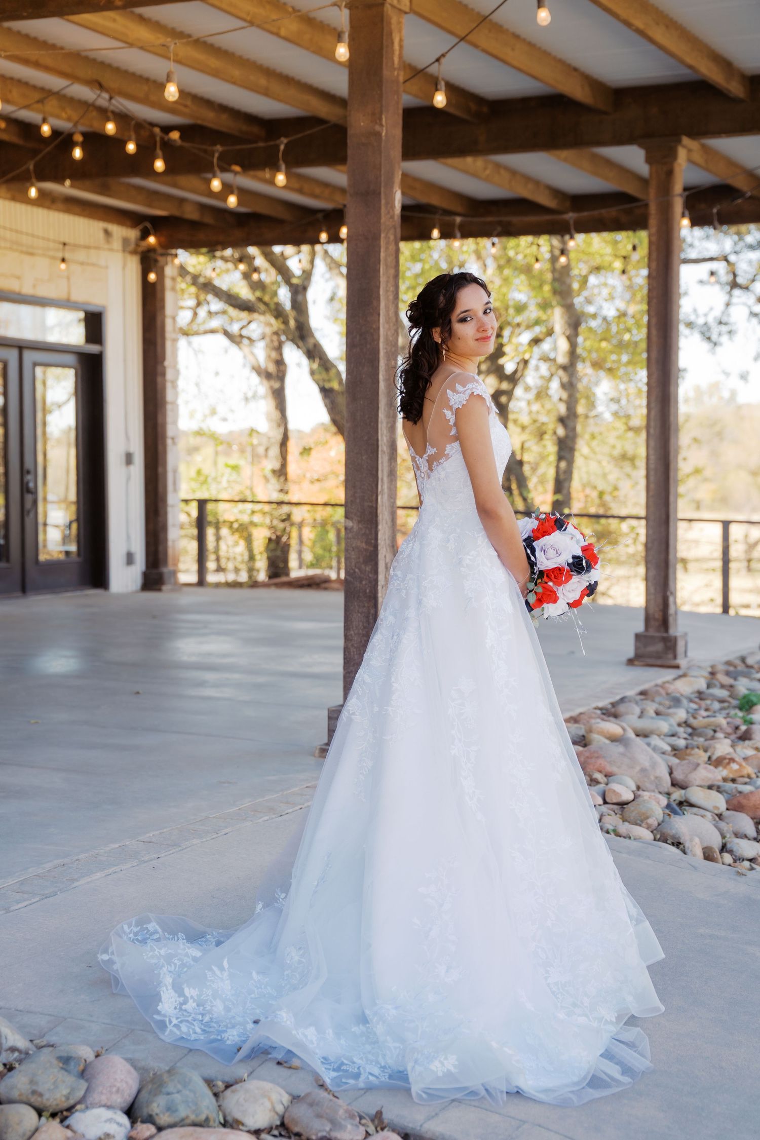 Bride in white lace ball gown holding red and blue bouquet poses on rustic venue patio with string lights. Oklahoma wedding photographer.