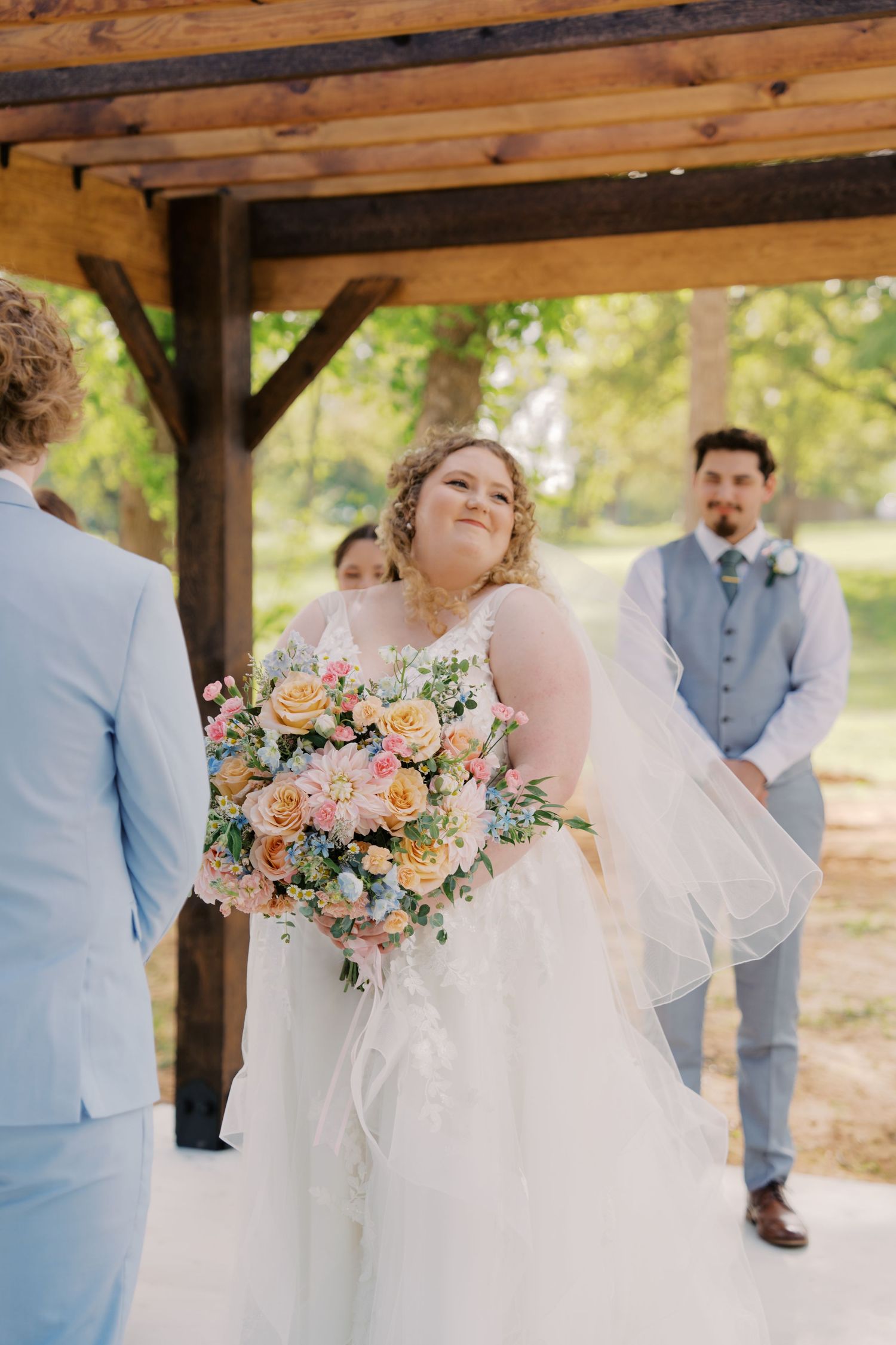 Bride in white gown holding colorful bouquet of roses smiles at groom during outdoor wedding ceremony under wooden pergola.