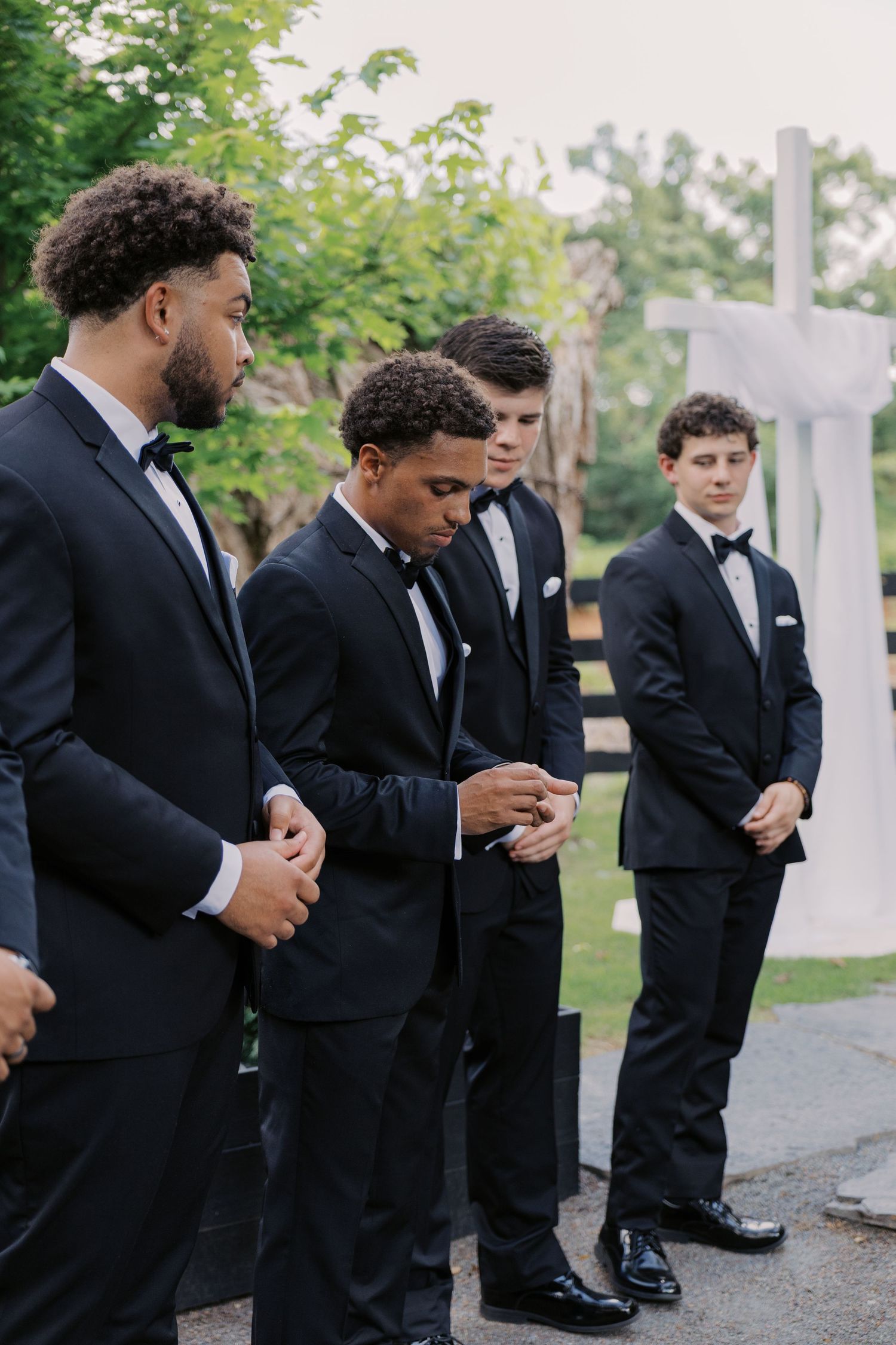Four groomsmen in black tuxedos bow their heads in prayer at an outdoor wedding ceremony. Oklahoma wedding photographer.