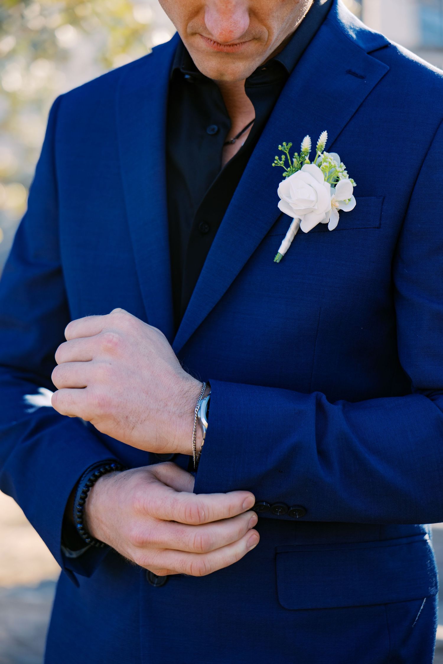 Groom in blue suit with white boutonniere adjusting his watch cufflink, wearing black shirt at wedding. Oklahoma wedding photographer.