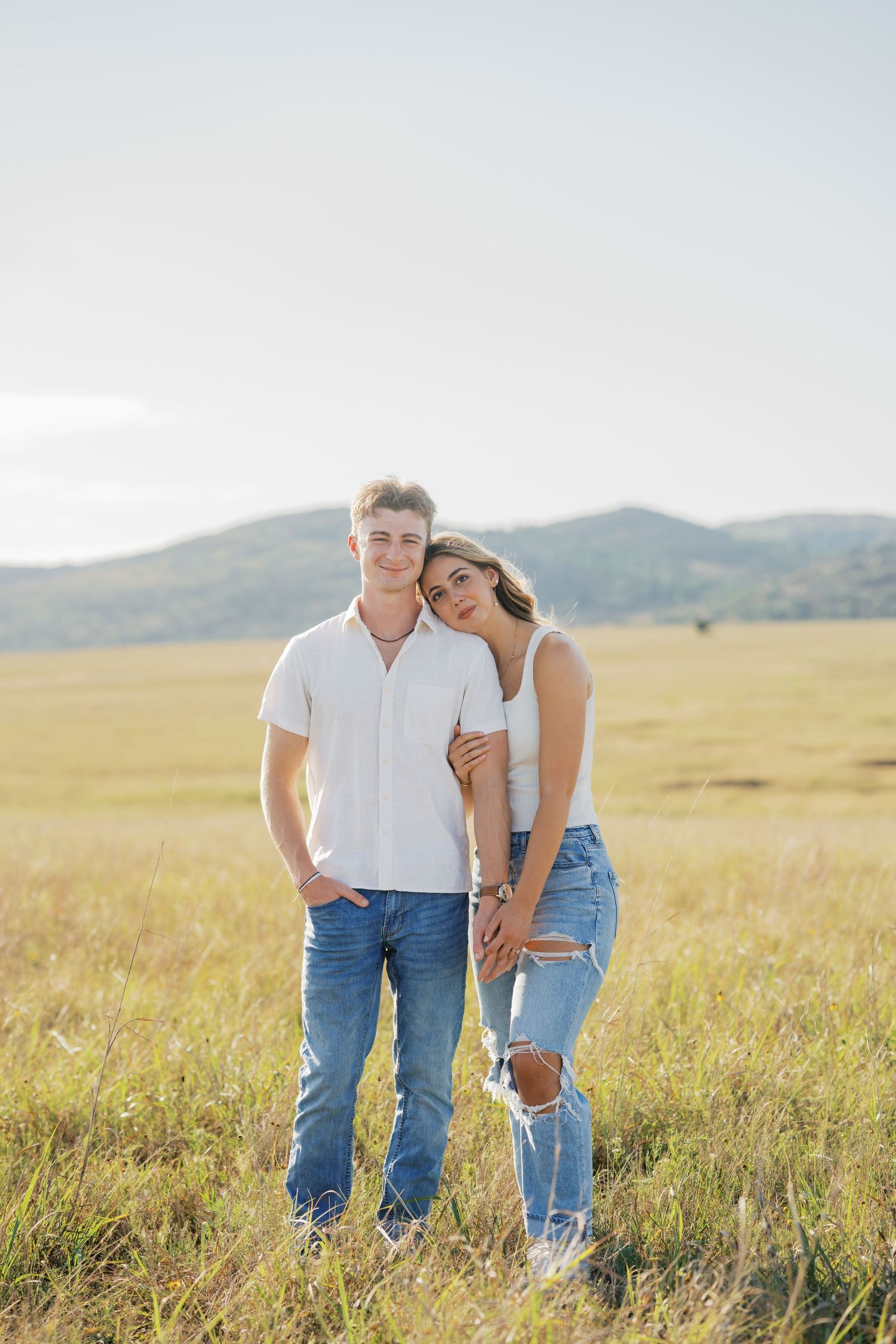 Young couple in white tops and jeans embracing in a golden field with mountain views.