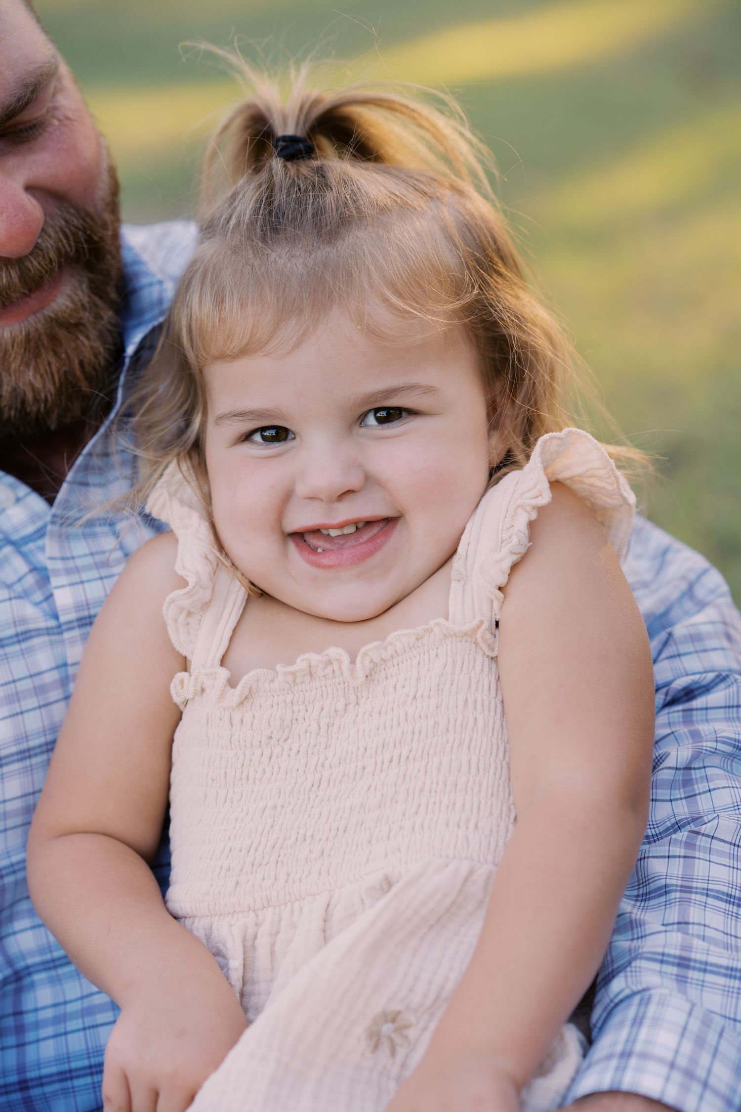 Smiling toddler girl with pigtails in a pink dress being held by a bearded man outdoors.