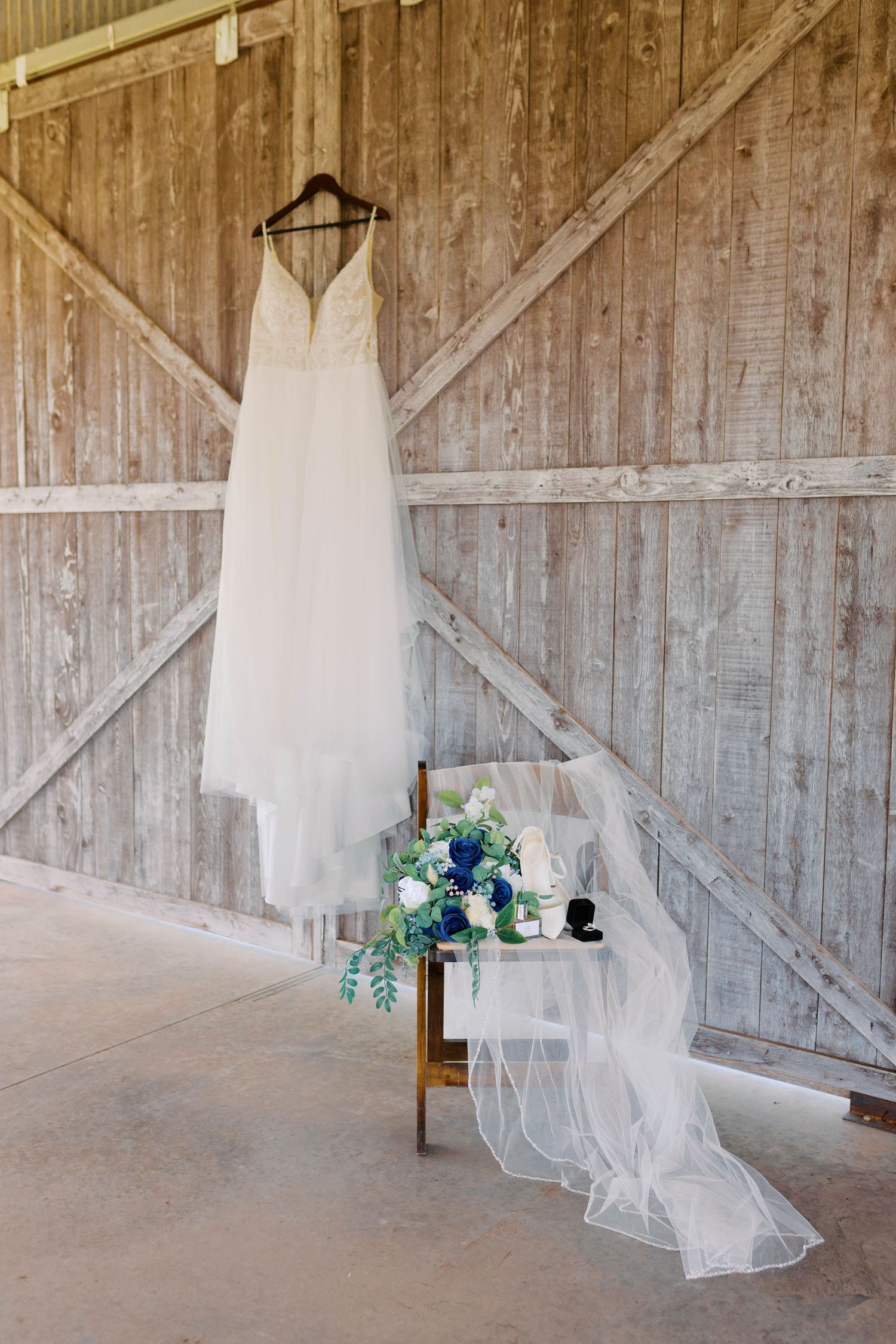 Elegant white wedding gown hanging in a rustic barn with a blue floral bouquet on a chair.