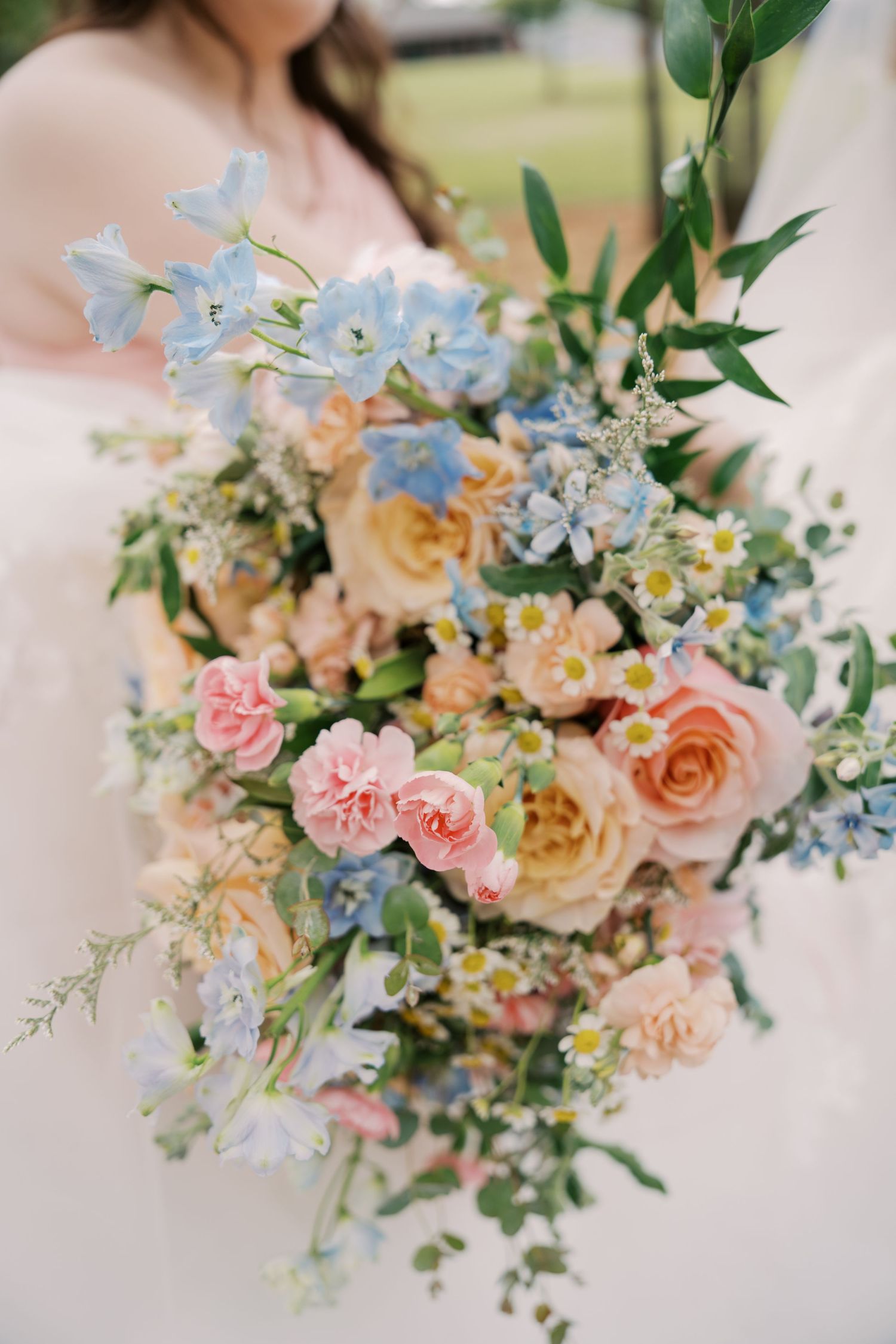 Bride holding a lush wedding bouquet with pink roses, blue delphiniums, peach blooms, daisies, and greenery. Oklahoma wedding photographer.