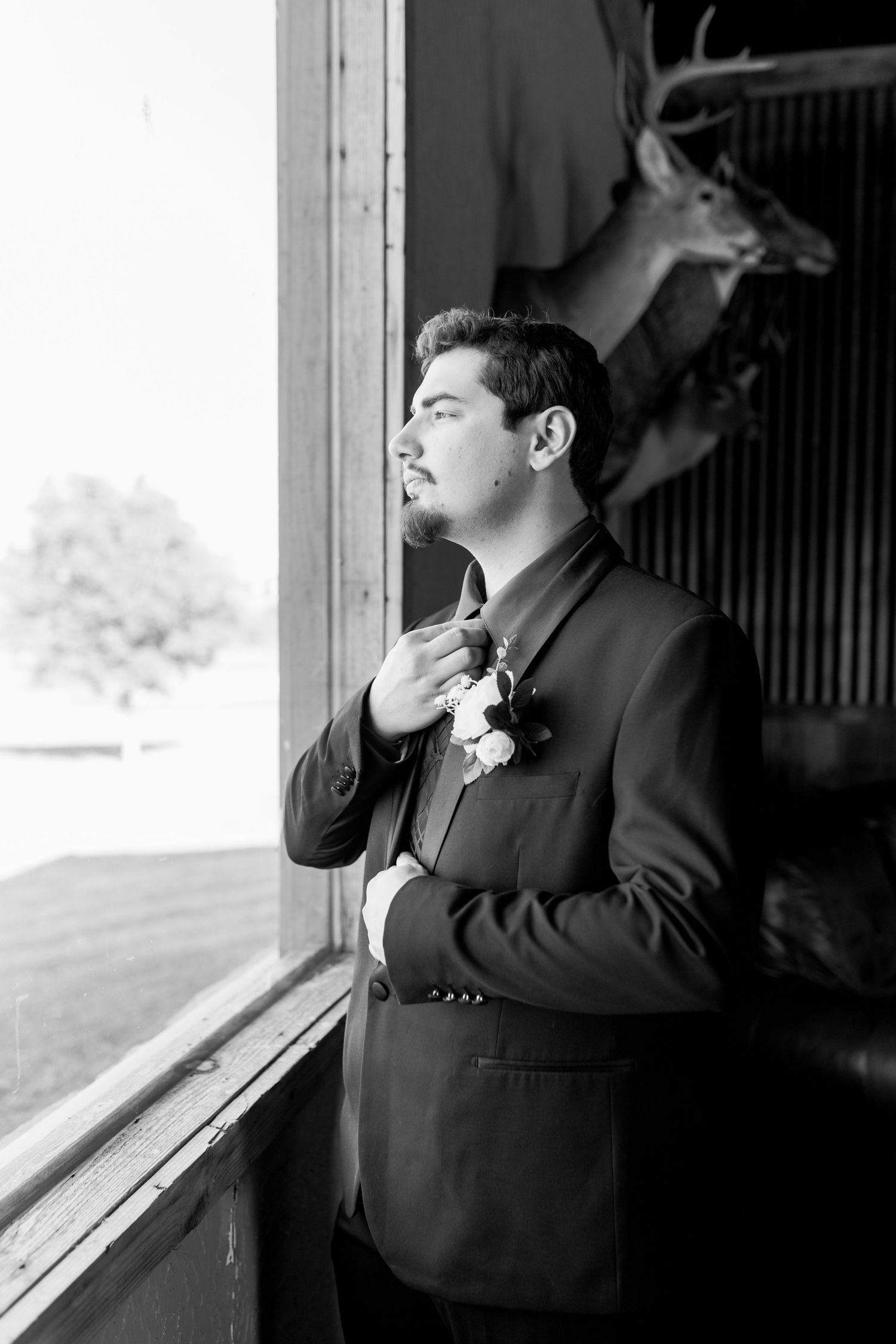 Groom in black suit adjusting his tie near a window, with a deer mount visible in the background. Oklahoma wedding photographer.