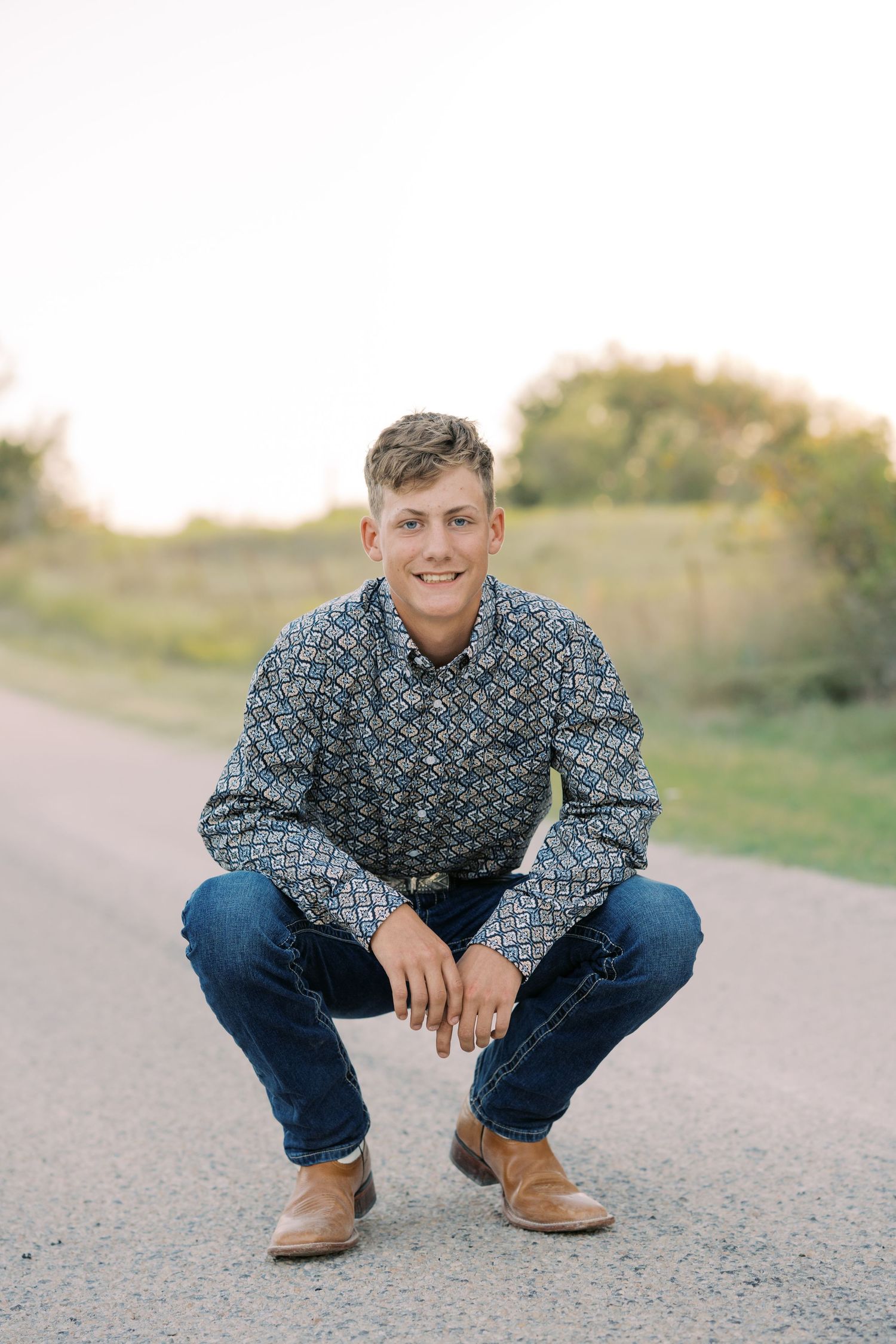 Young man in patterned shirt and jeans crouching on a rural road, smiling confidently during golden hour.