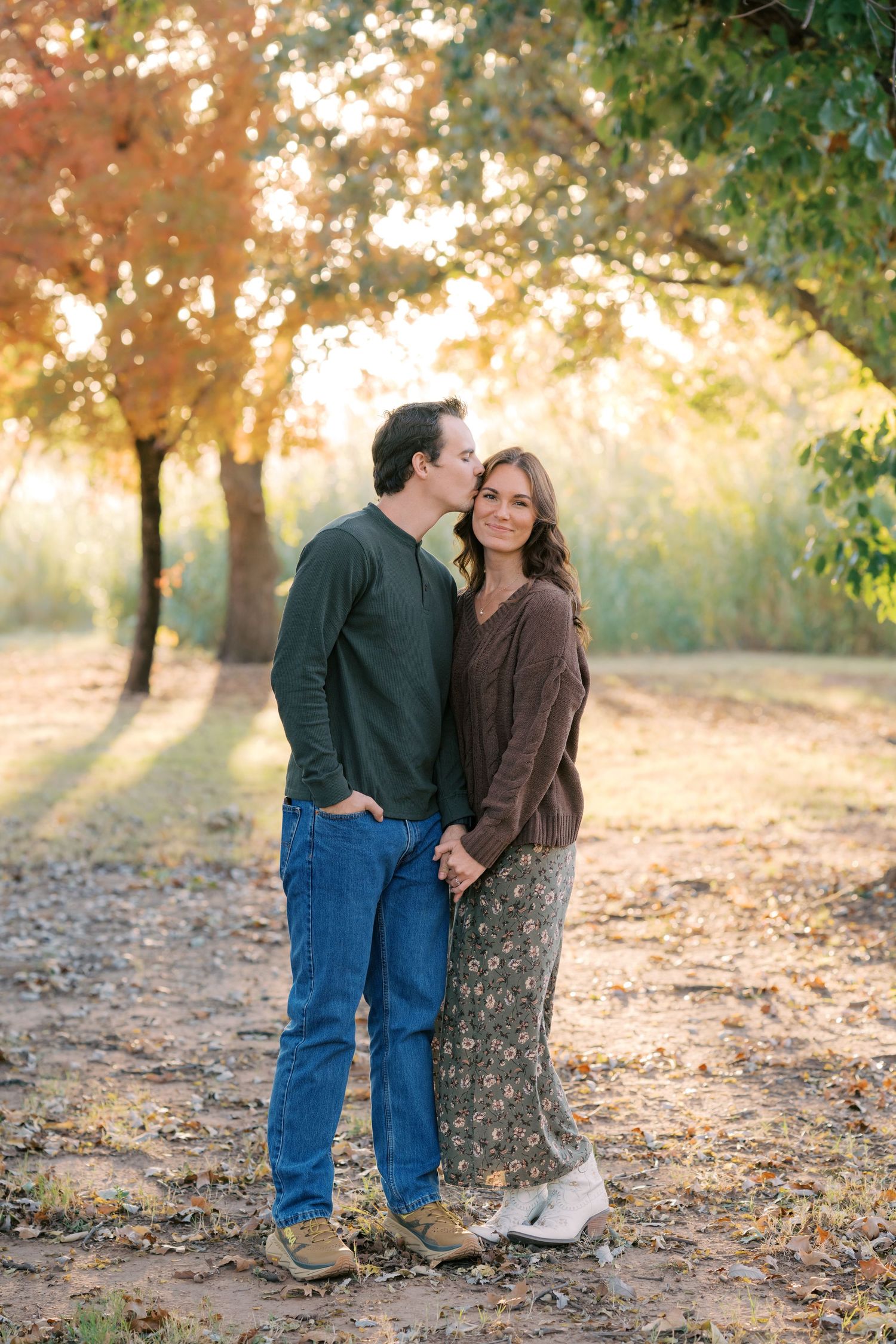 A couple holds hands and shares a kiss among autumn trees with golden foliage in a romantic outdoor portrait.