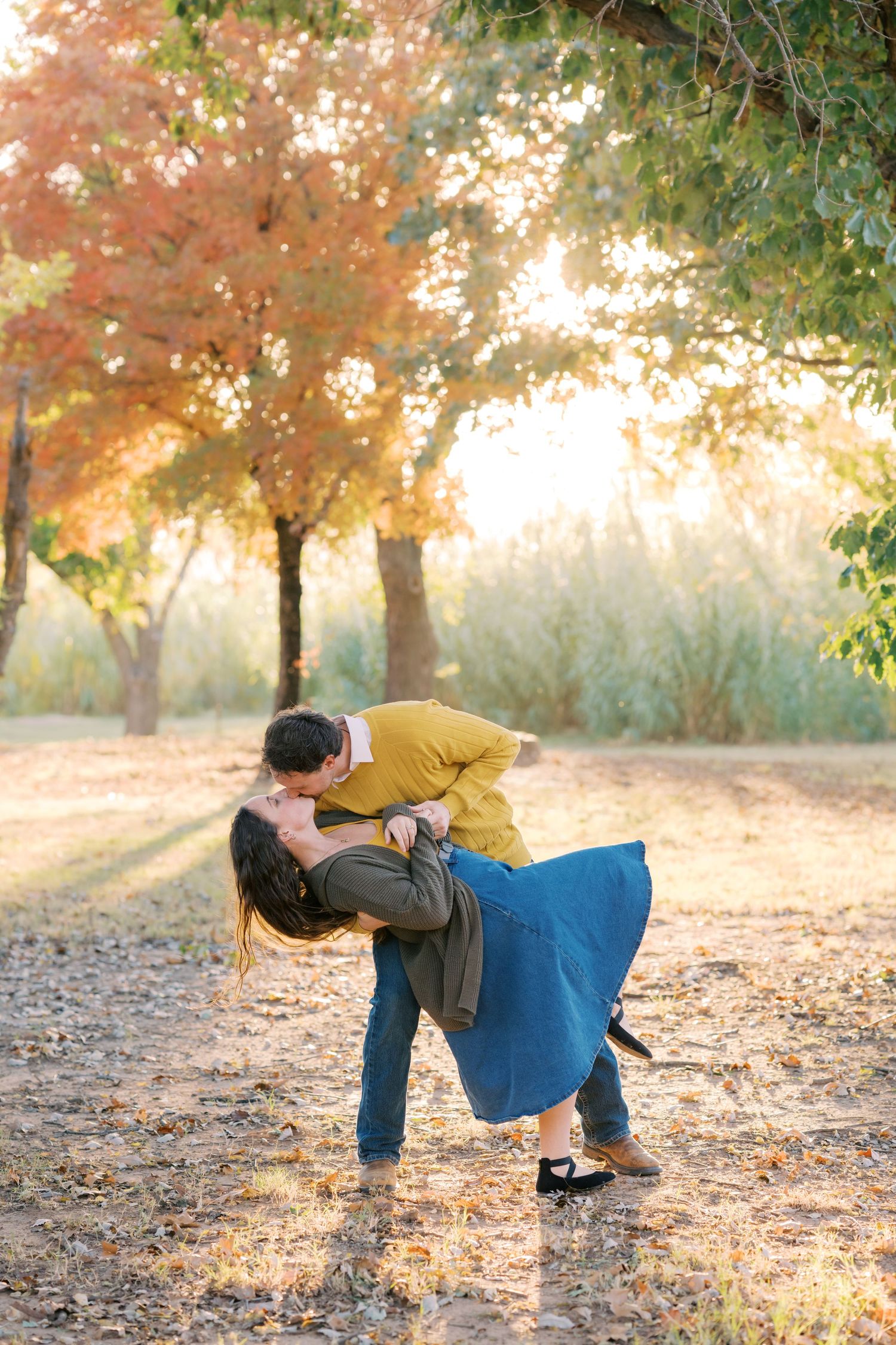 Couple sharing a romantic dip kiss surrounded by colorful autumn trees in golden sunlight.