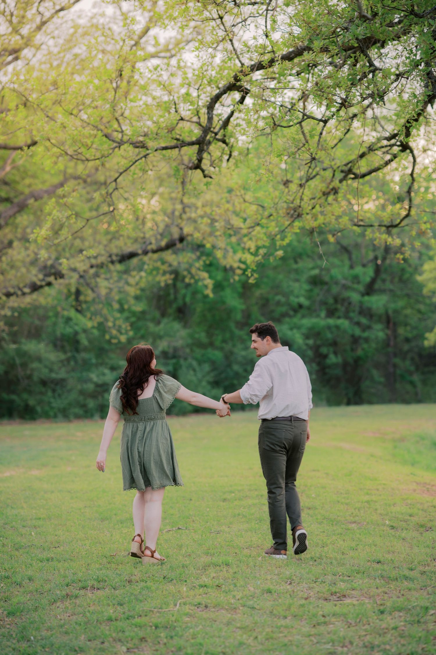 A couple holds hands while walking through a lush green park, the woman in a sage dress and man in a white shirt.