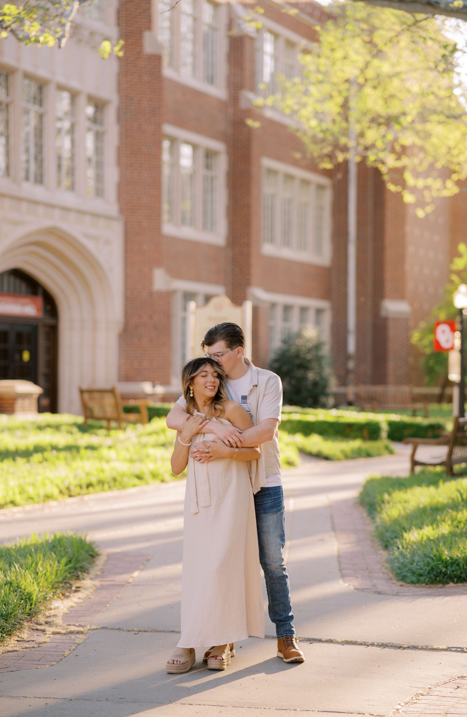 A couple embraces on a sunny campus walkway in front of a classic brick university building.