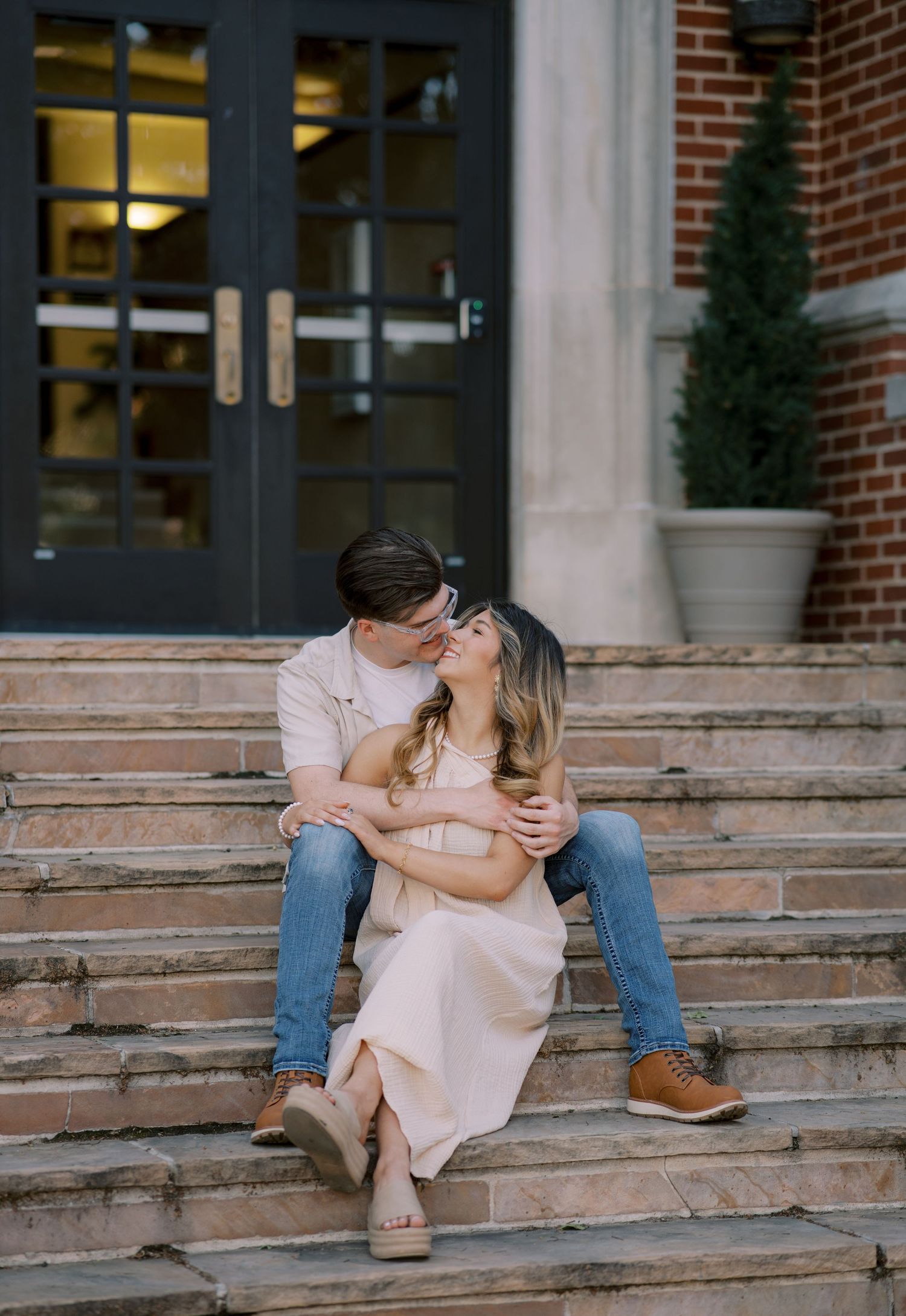 Couple sitting on brick steps sharing a romantic kiss, woman in blush dress, man in jeans embracing her tenderly. Oklahoma wedding.