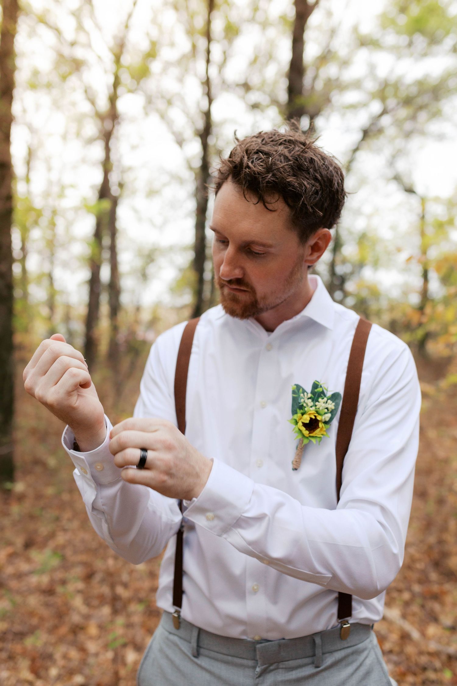 Groom in white shirt with brown suspenders and sunflower boutonniere adjusts cufflinks in autumn forest. Oklahoma wedding photographer.