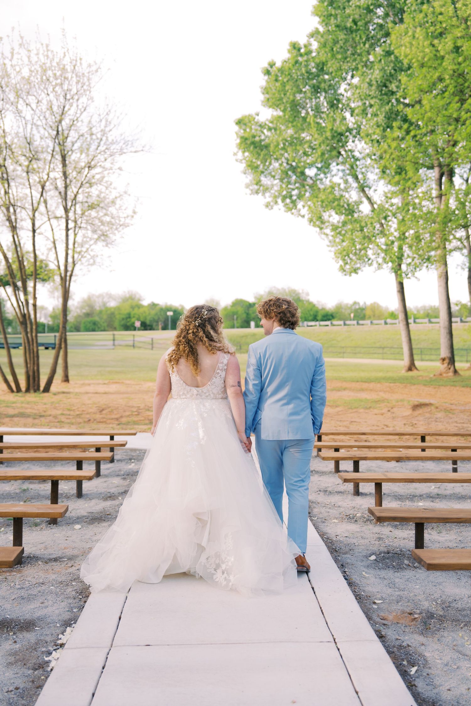 Bride in white gown and groom in light blue suit walk hand in hand down aisle after outdoor ceremony. Oklahoma wedding photographer.