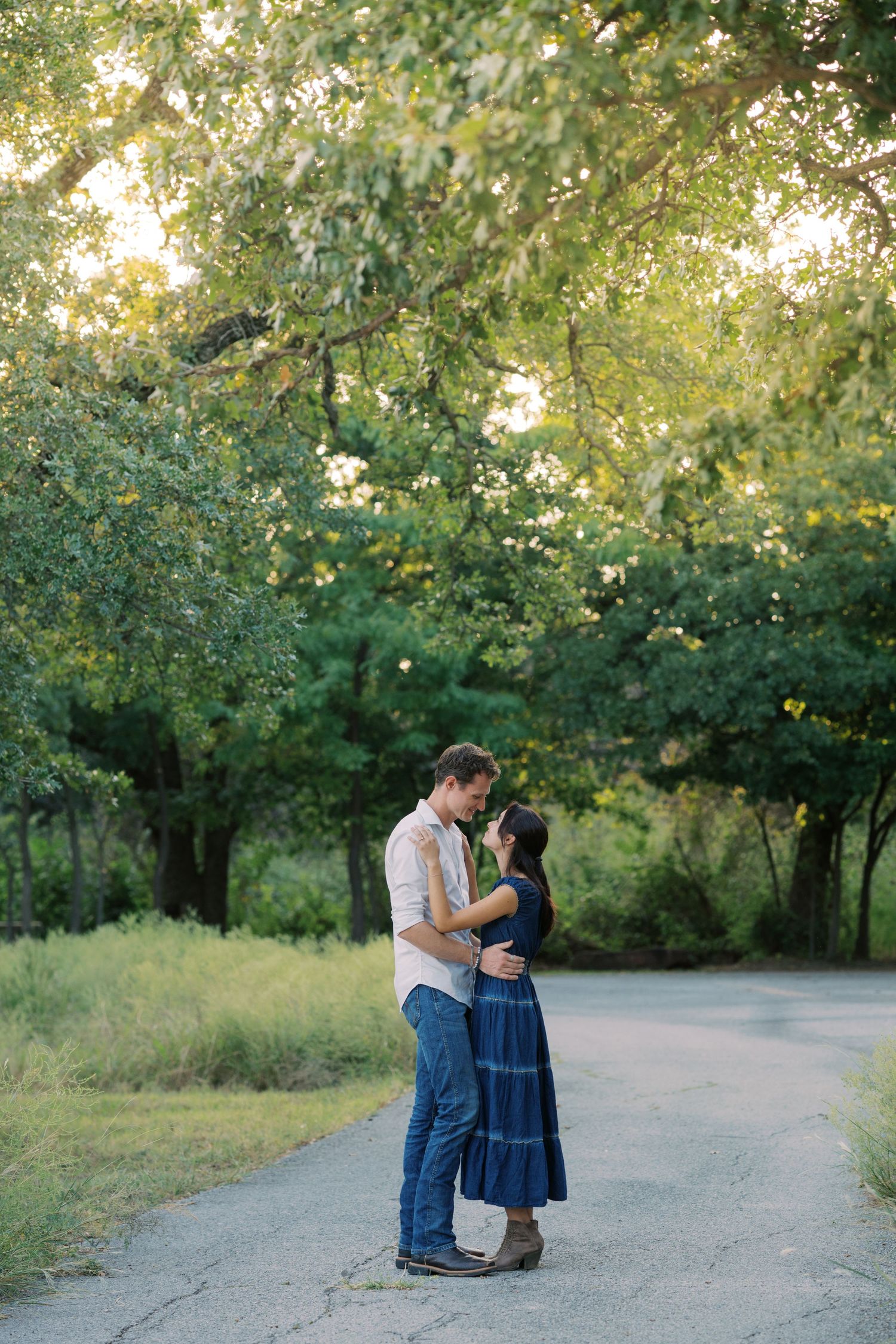 Couple embracing on a tree-lined path during golden hour, woman in blue dress and man in jeans sharing a romantic moment.