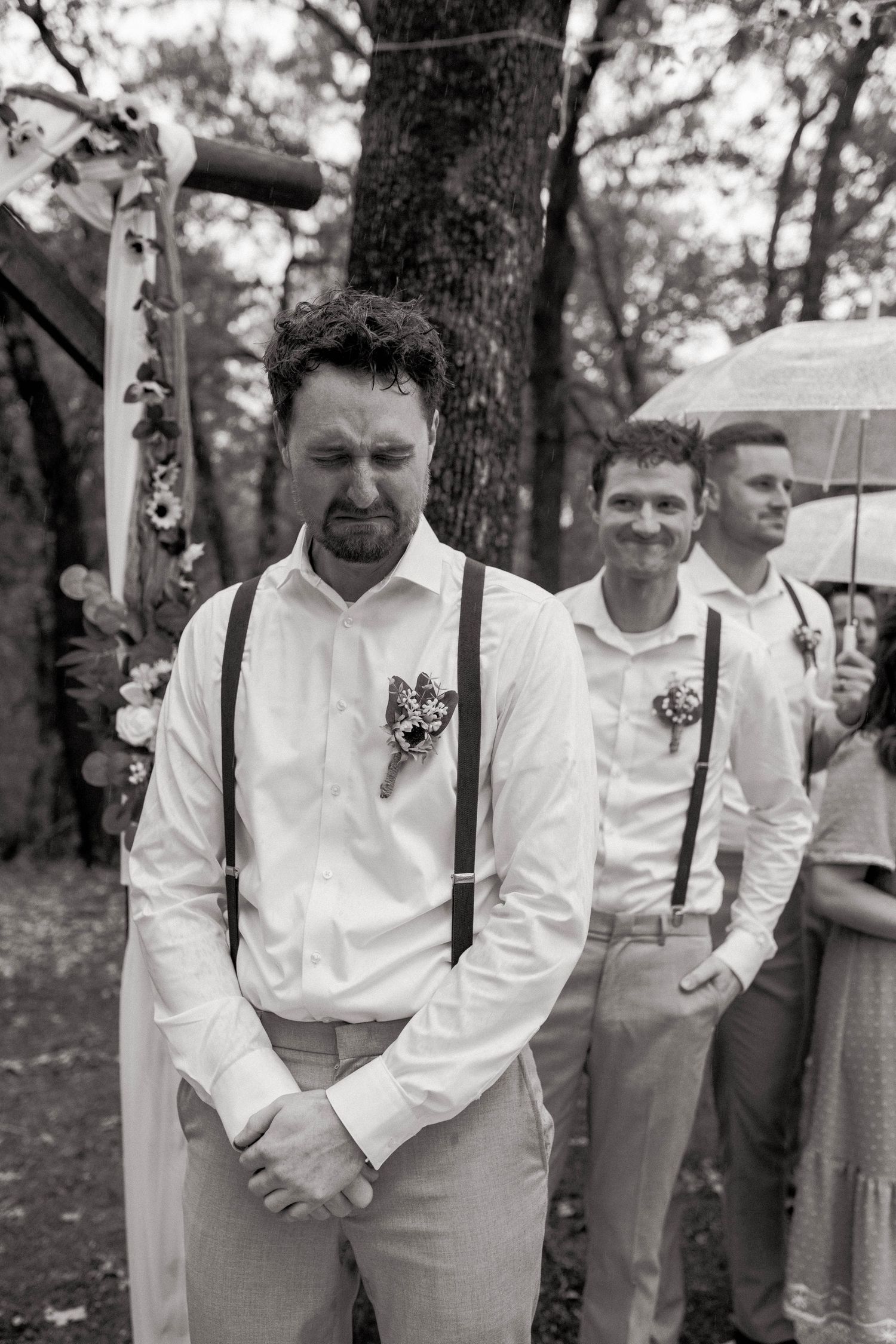Emotional groom in white shirt and suspenders stands at outdoor wedding altar in black and white photo. Oklahoma wedding photographer.