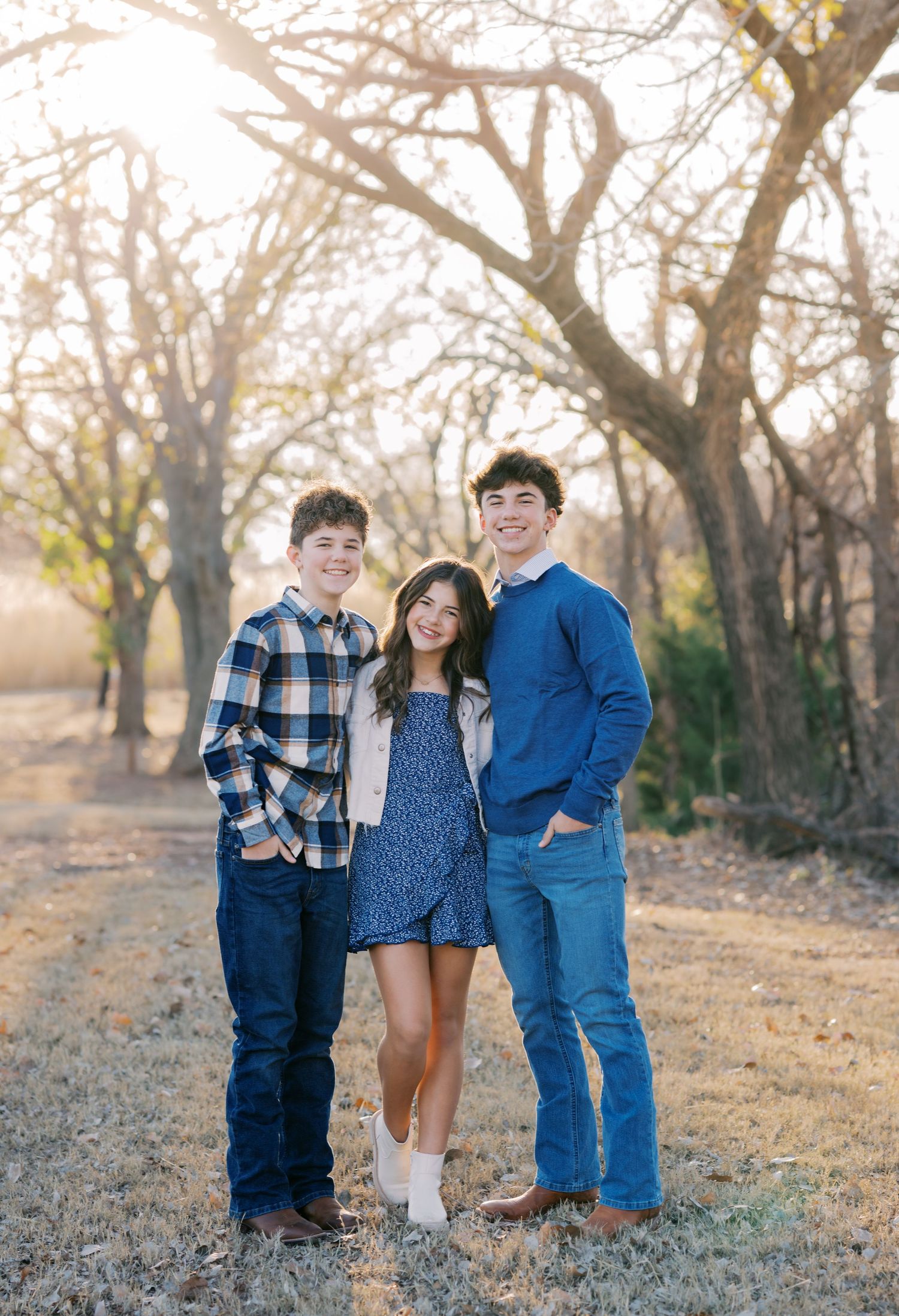 Three smiling teenagers pose together outdoors during golden hour among bare trees in a sunny field.