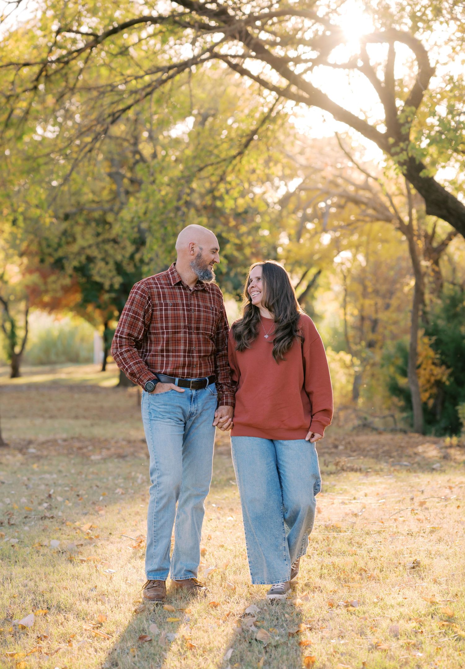 Couple holding hands and smiling at each other while walking through a sunlit autumn park with golden foliage.