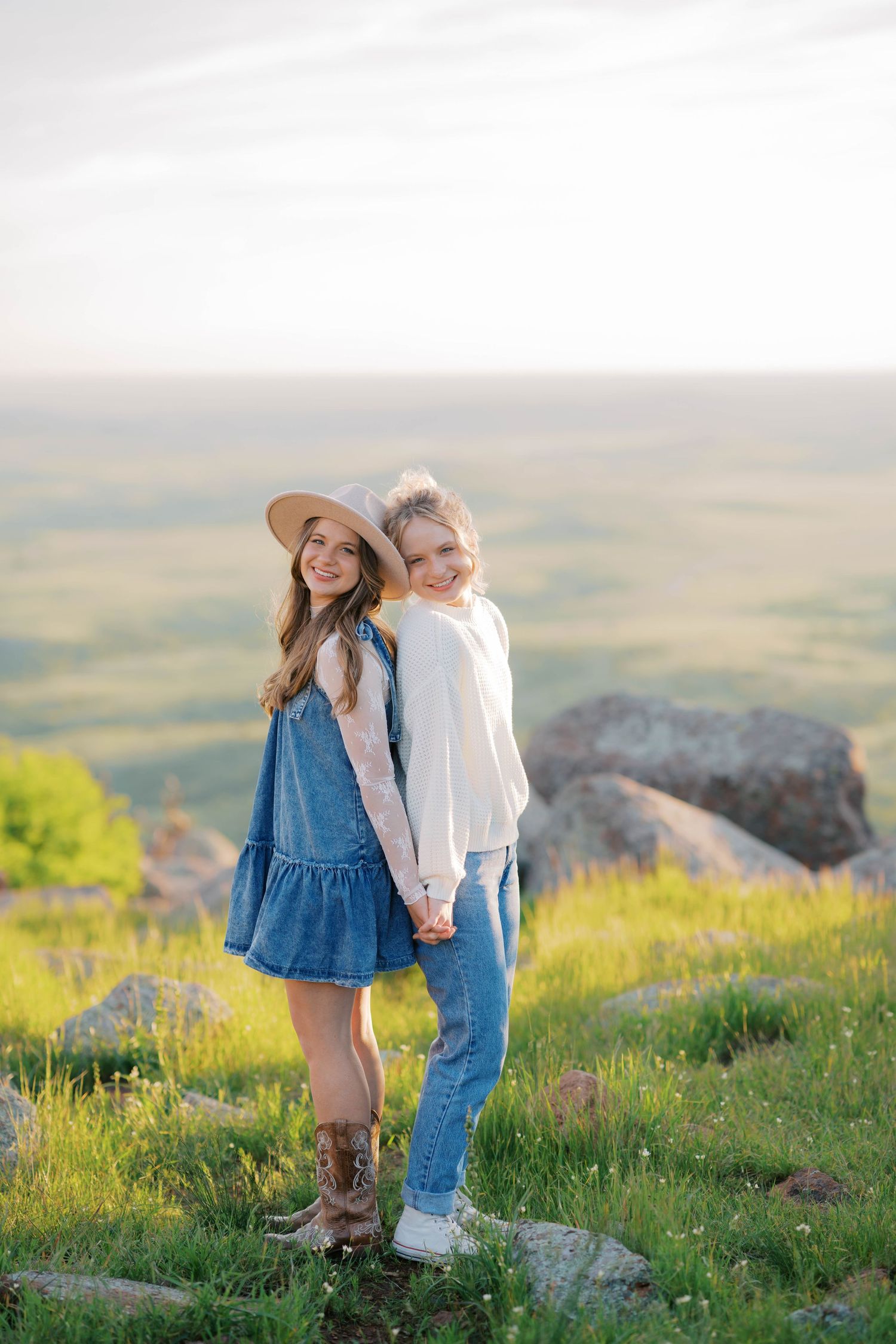 Two young women stand back-to-back holding hands on a scenic hilltop, one wearing a denim dress and hat, the other in jeans.