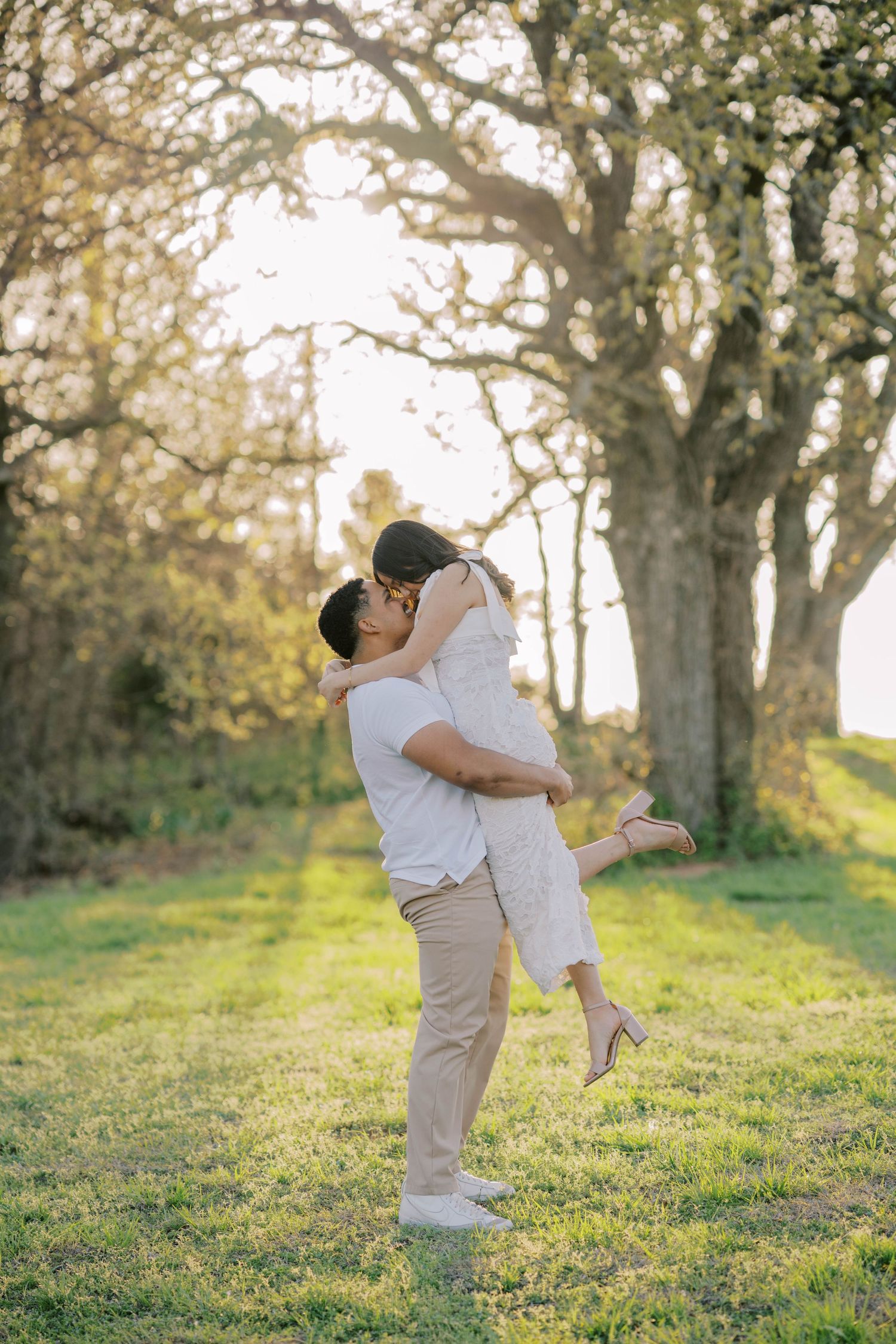 Man lifts and kisses woman in a sunlit park during a romantic outdoor engagement photo session.