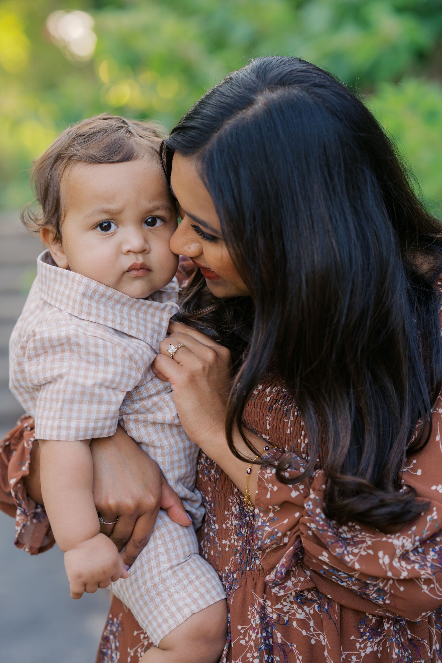 A loving mother with dark hair in a floral dress holds and kisses her baby boy outdoors in a lush green setting.