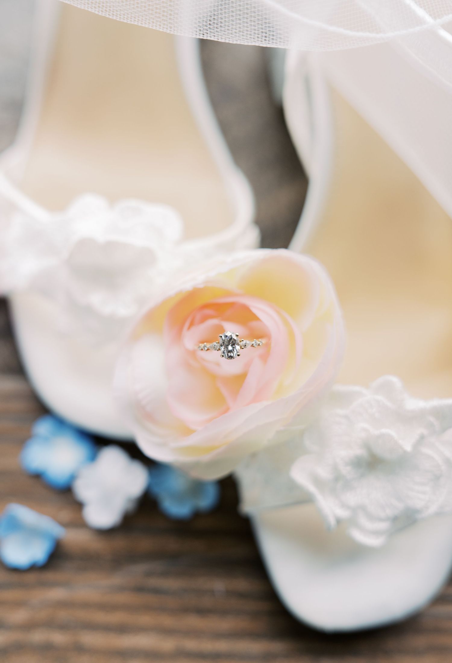 Elegant bridal shoes adorned with silk roses displaying a diamond engagement ring, surrounded by blue petals.