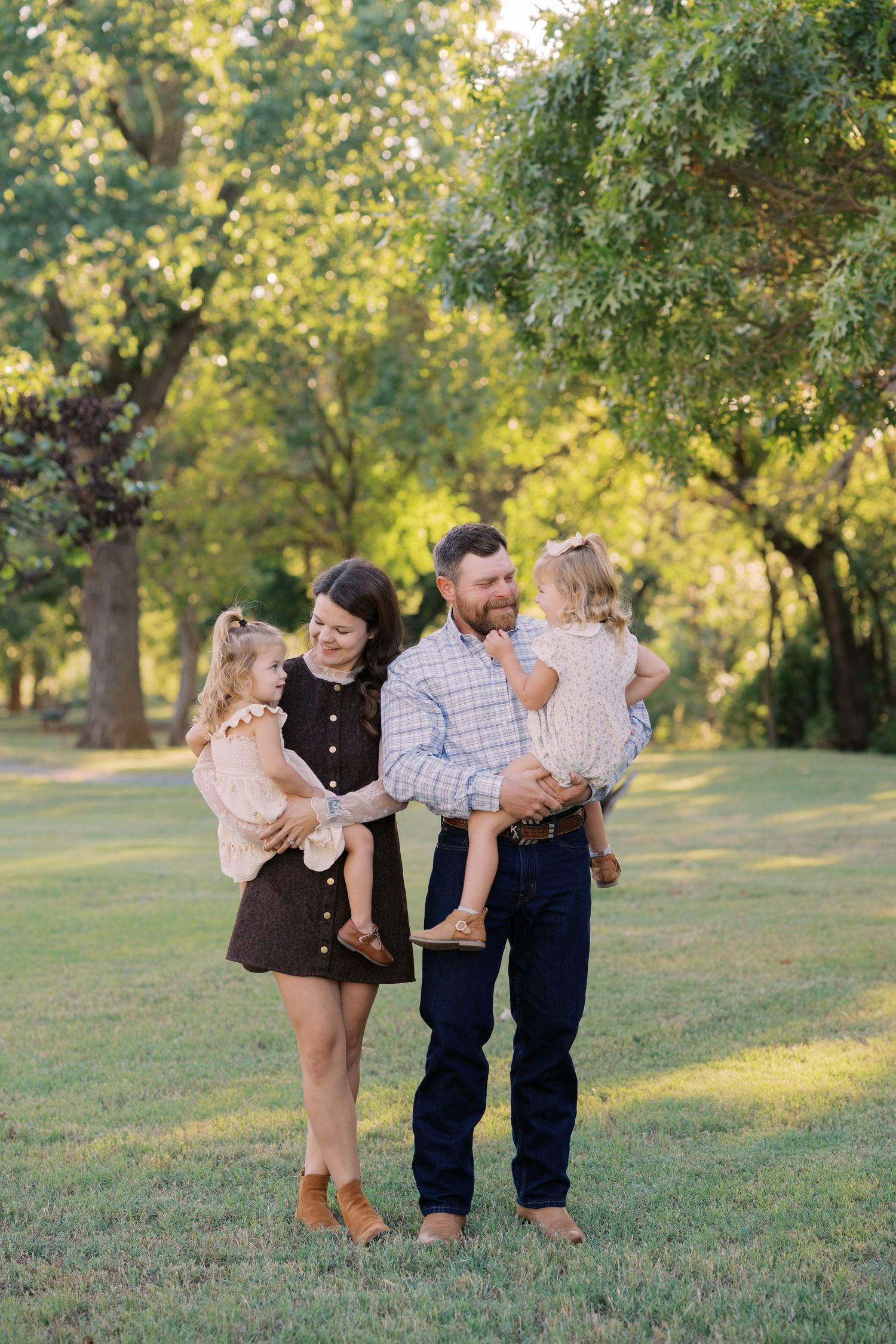 A happy family of four poses outdoors in a lush green park, parents holding two young daughters during golden hour.