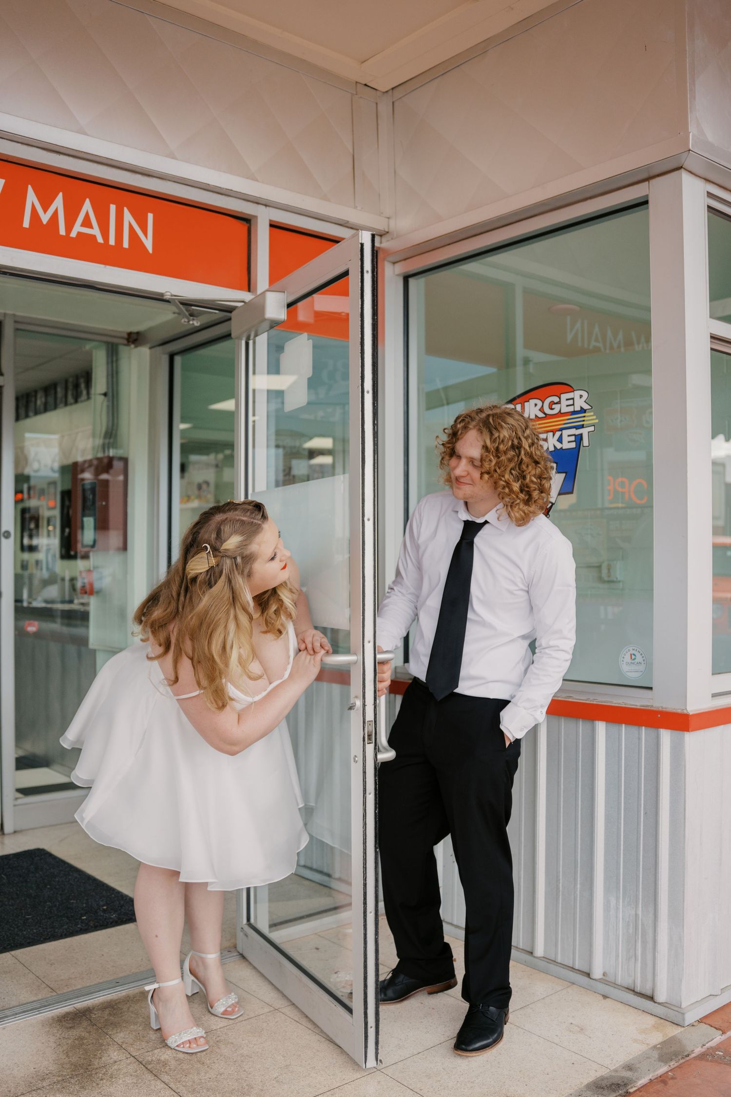 A bride in a short white dress and groom in a black tie stand at a storefront entrance.
