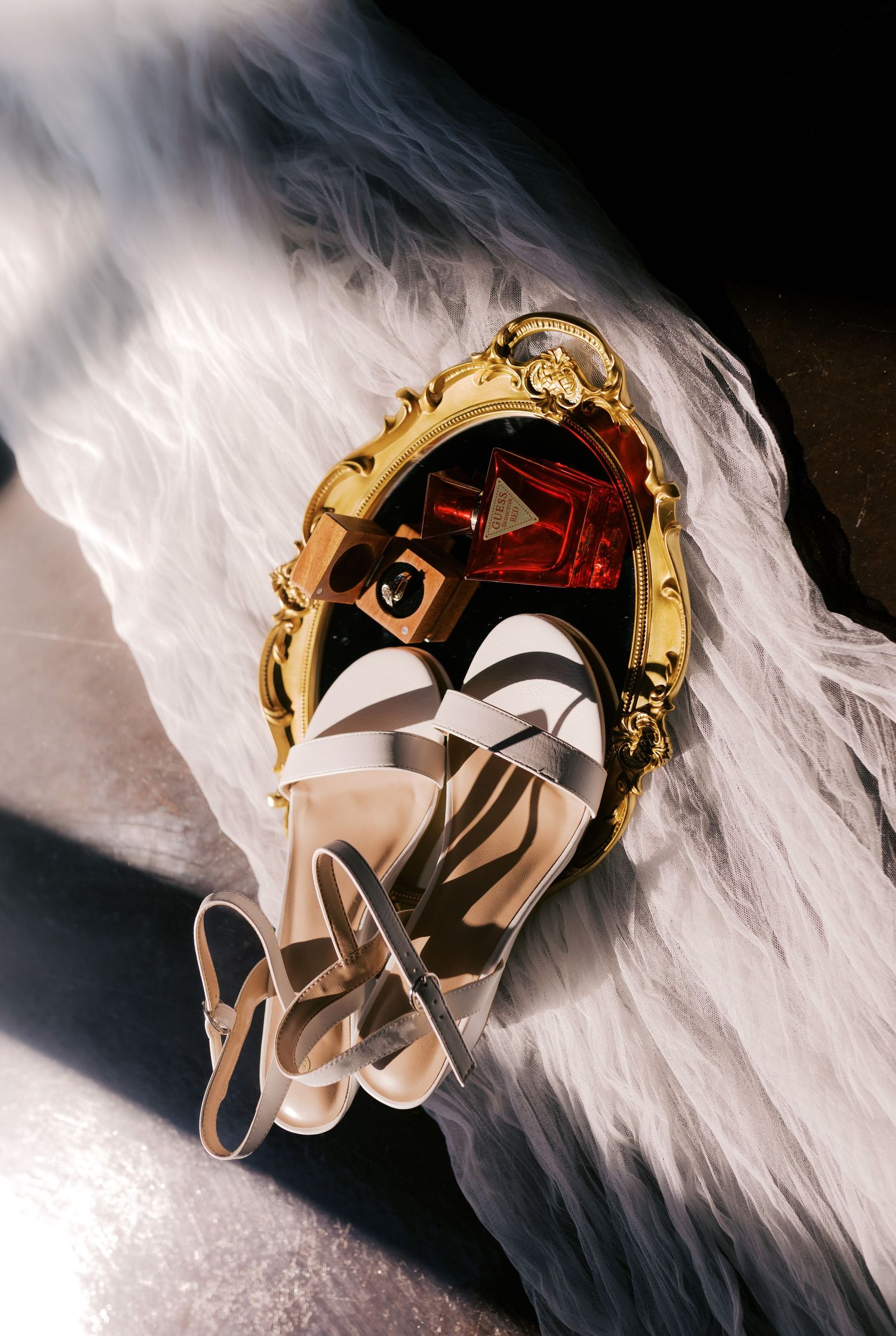 Gold ornate mirror with white tulle, red perfume bottle, and white strappy sandals in a dramatic flat lay arrangement.