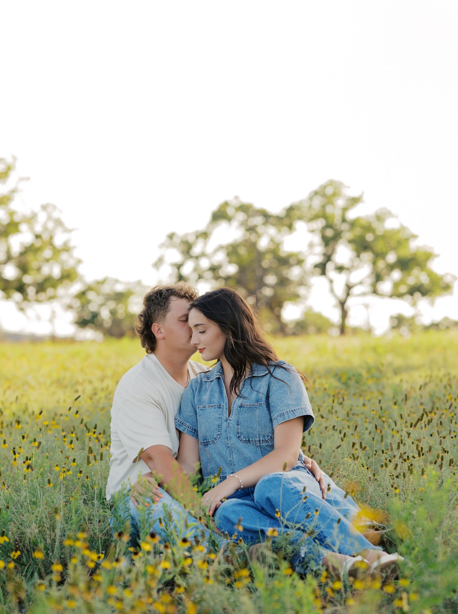 Couple sitting in a wildflower field, sharing a tender kiss, surrounded by lush greenery and trees in soft golden light.