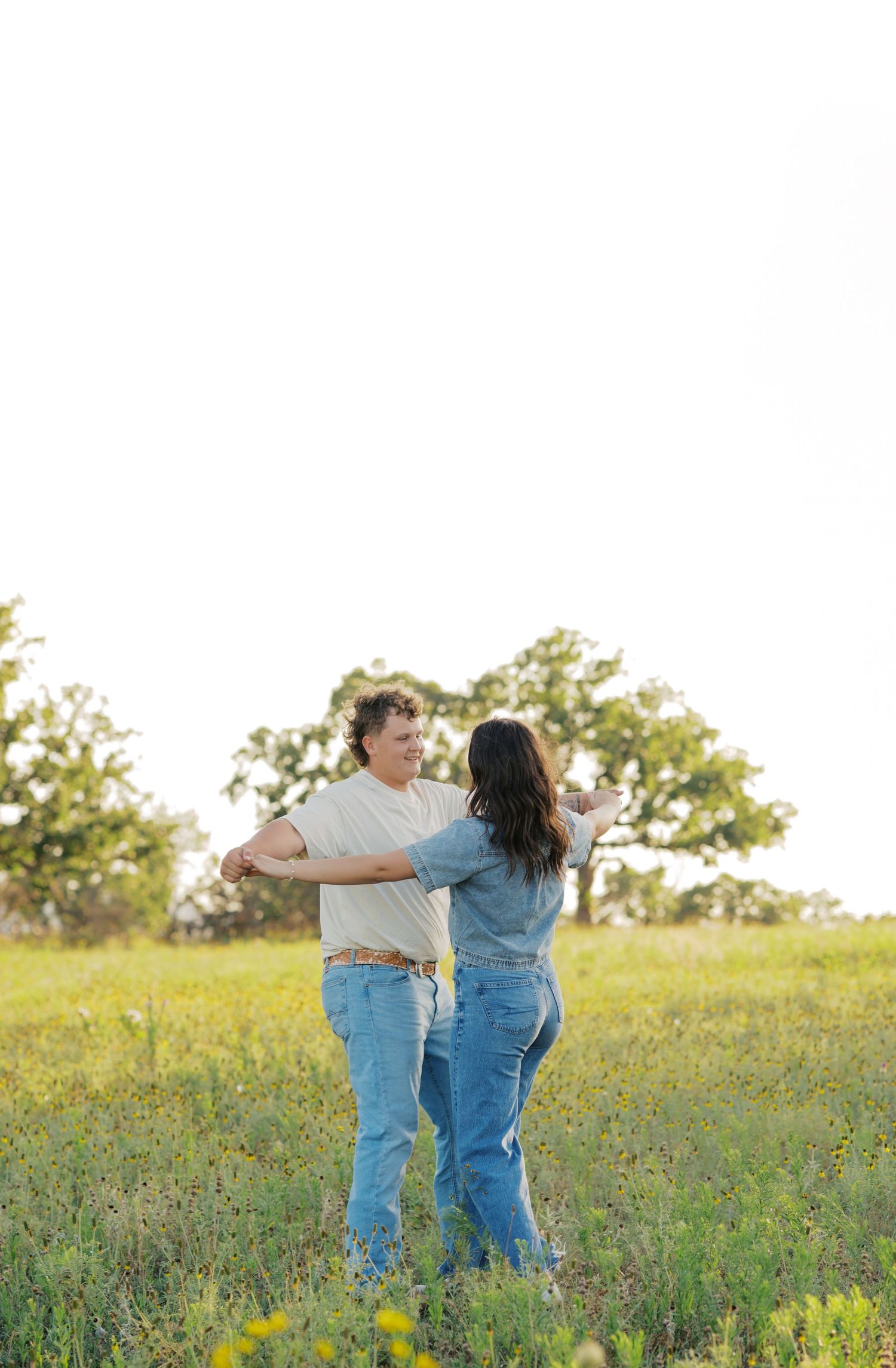 Couple dancing together in a sunny green meadow, wearing jeans and casual tops, with trees in the background.