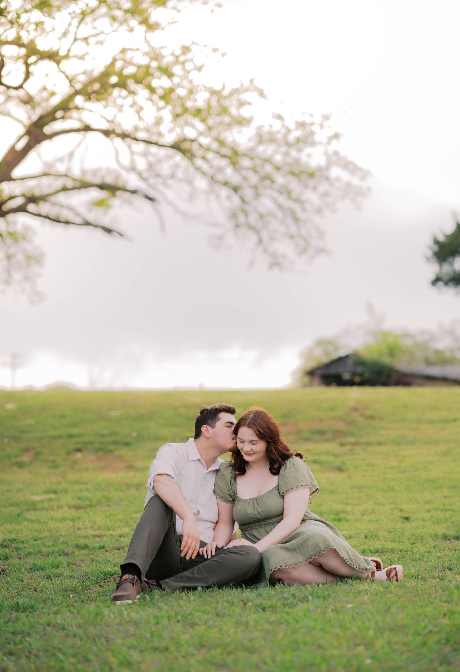 Couple sits on grass sharing a kiss beneath a tree during a romantic golden hour outdoor session.