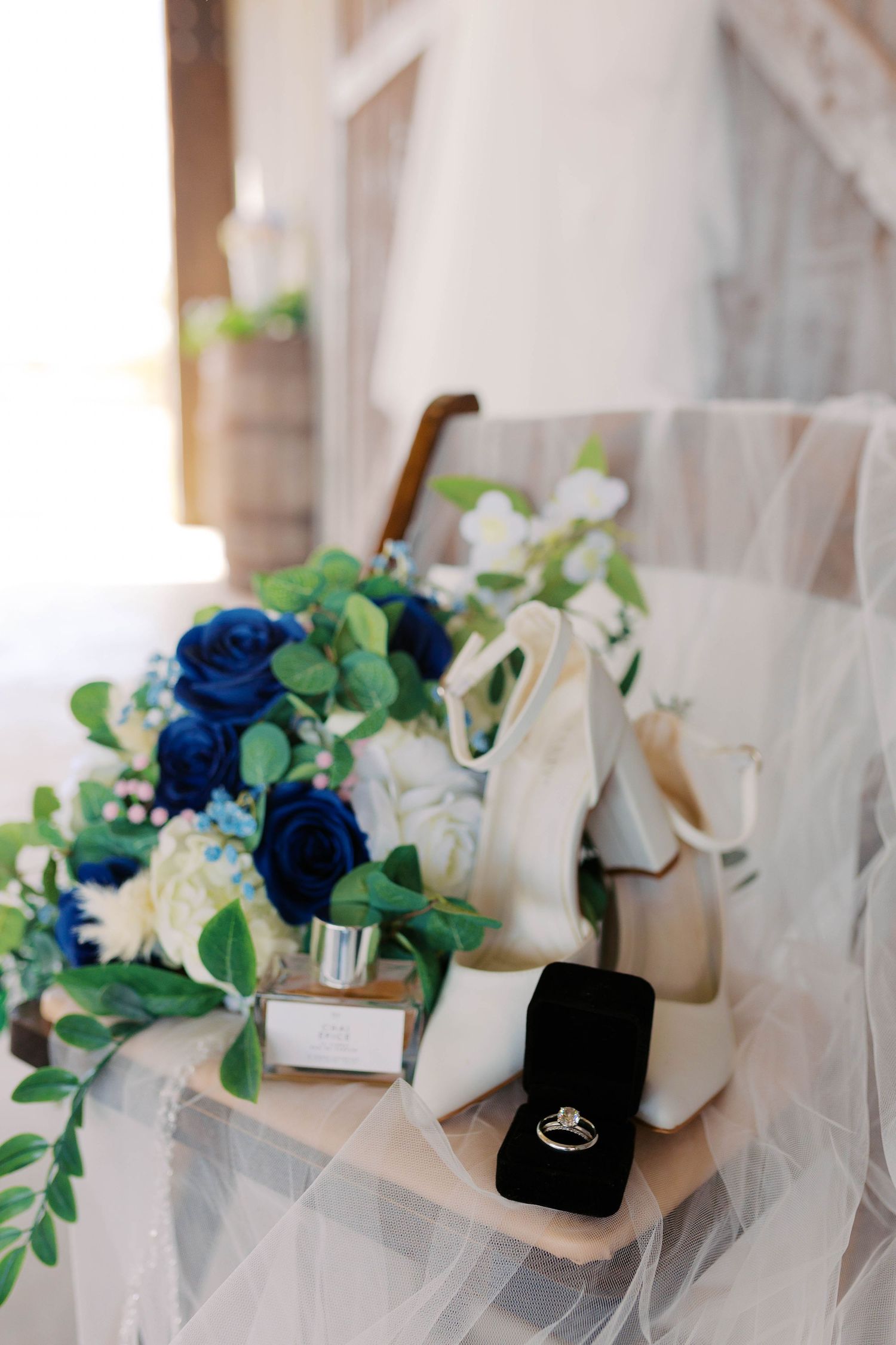 Wedding flat lay featuring blue rose bouquet, white heels, perfume, and diamond ring in velvet box.