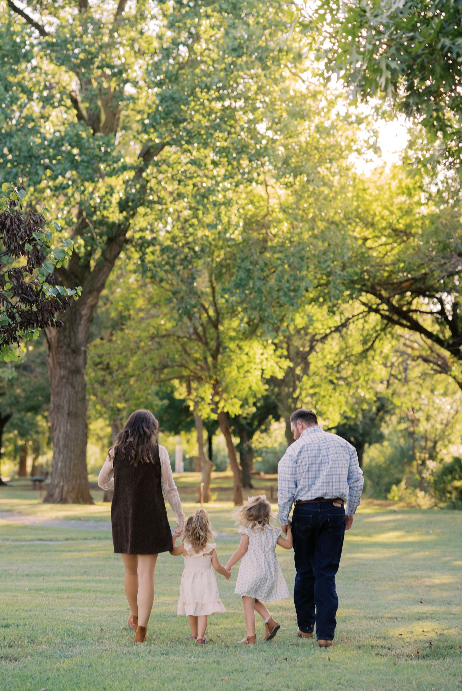 A family of four walks hand-in-hand through a sunlit park, with two young girls in white dresses between their parents.