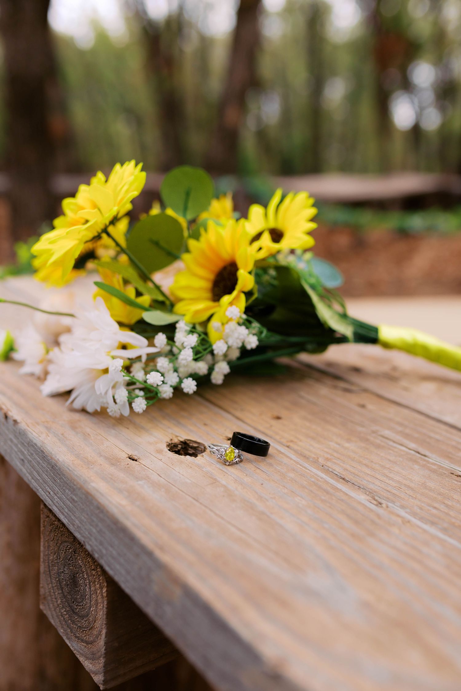 Wedding rings and a bridal bouquet of sunflowers and white flowers rest on a wooden surface.