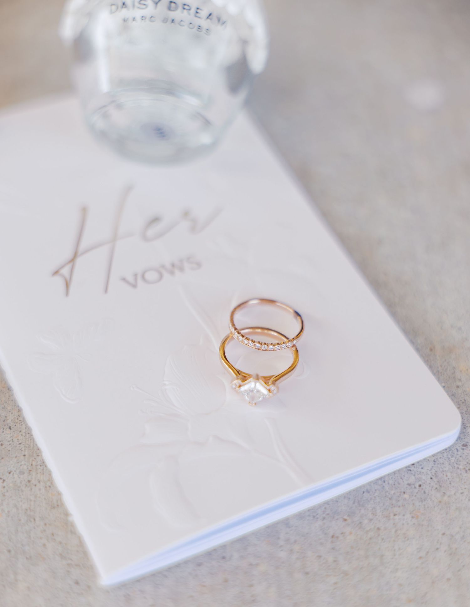 Gold engagement ring and wedding band resting on a white Her Vows book with a perfume bottle in the background.