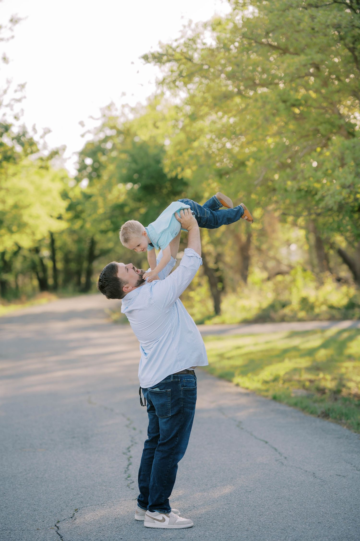 A father joyfully lifts his baby high in the air on a sunny tree-lined path outdoors.
