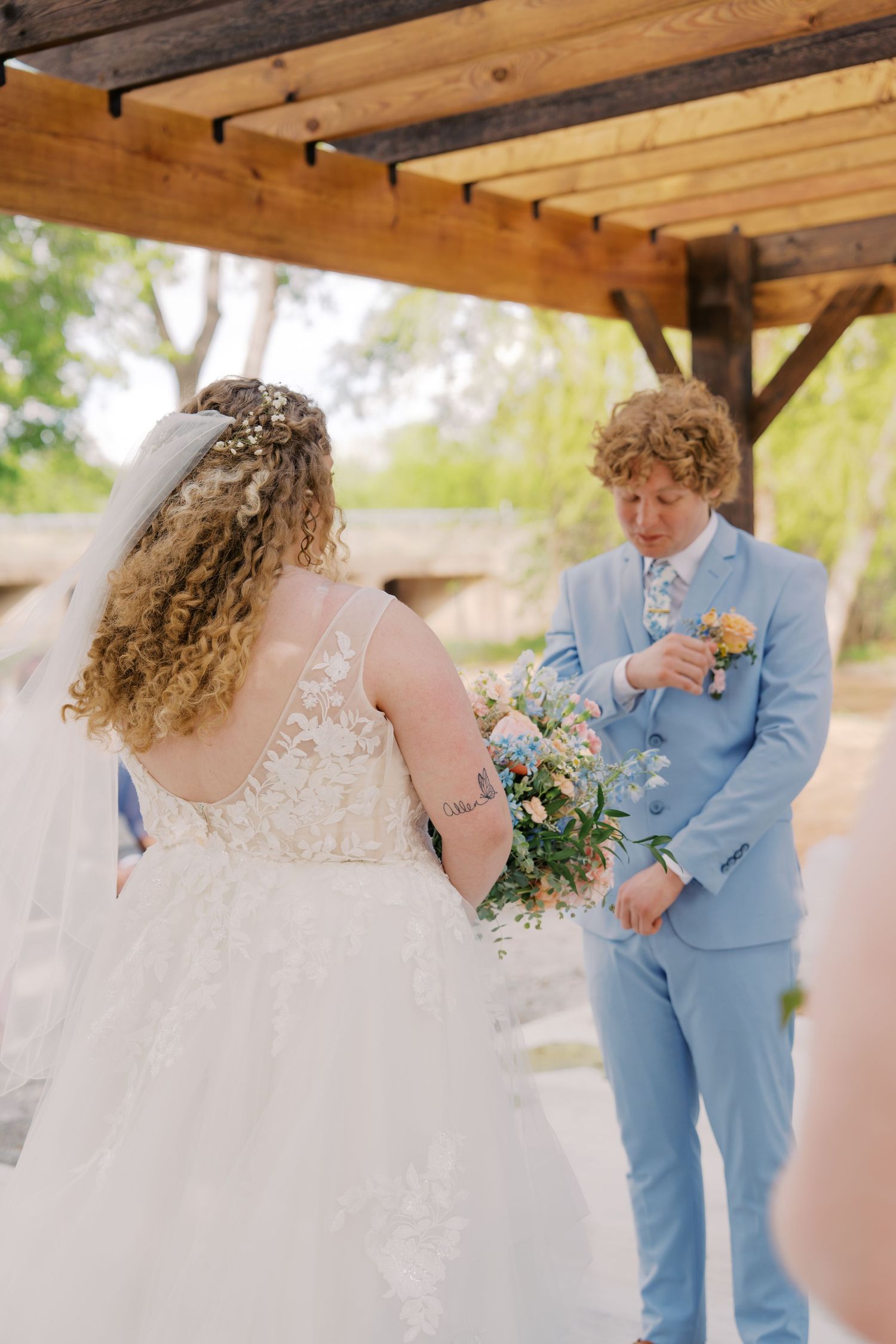 Bride in lace gown holding wildflower bouquet faces groom in light blue suit at outdoor wedding ceremony.