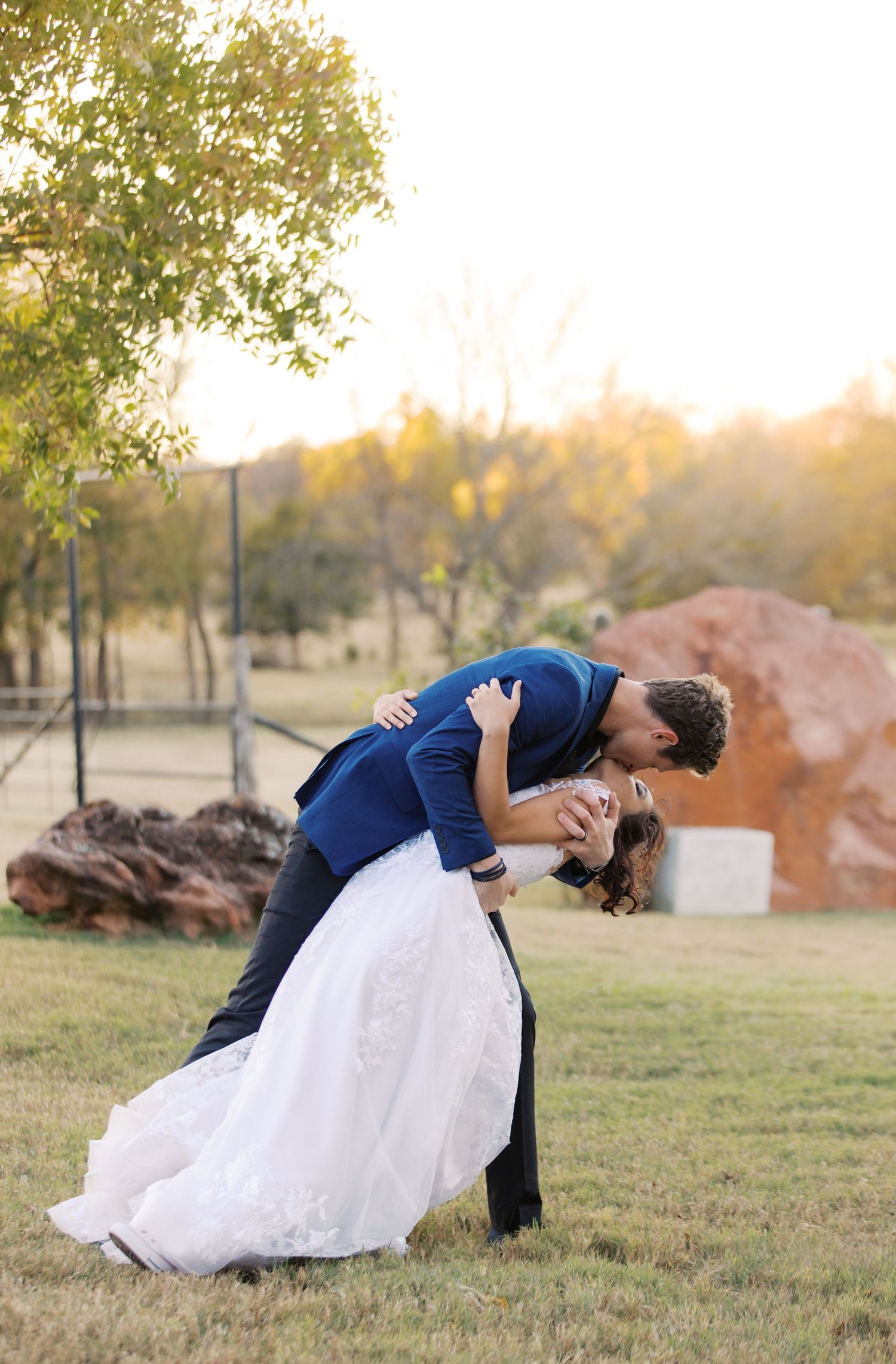 Groom in navy suit dips bride in white lace gown for a romantic kiss outdoors at golden hour. Oklahoma wedding photographer.