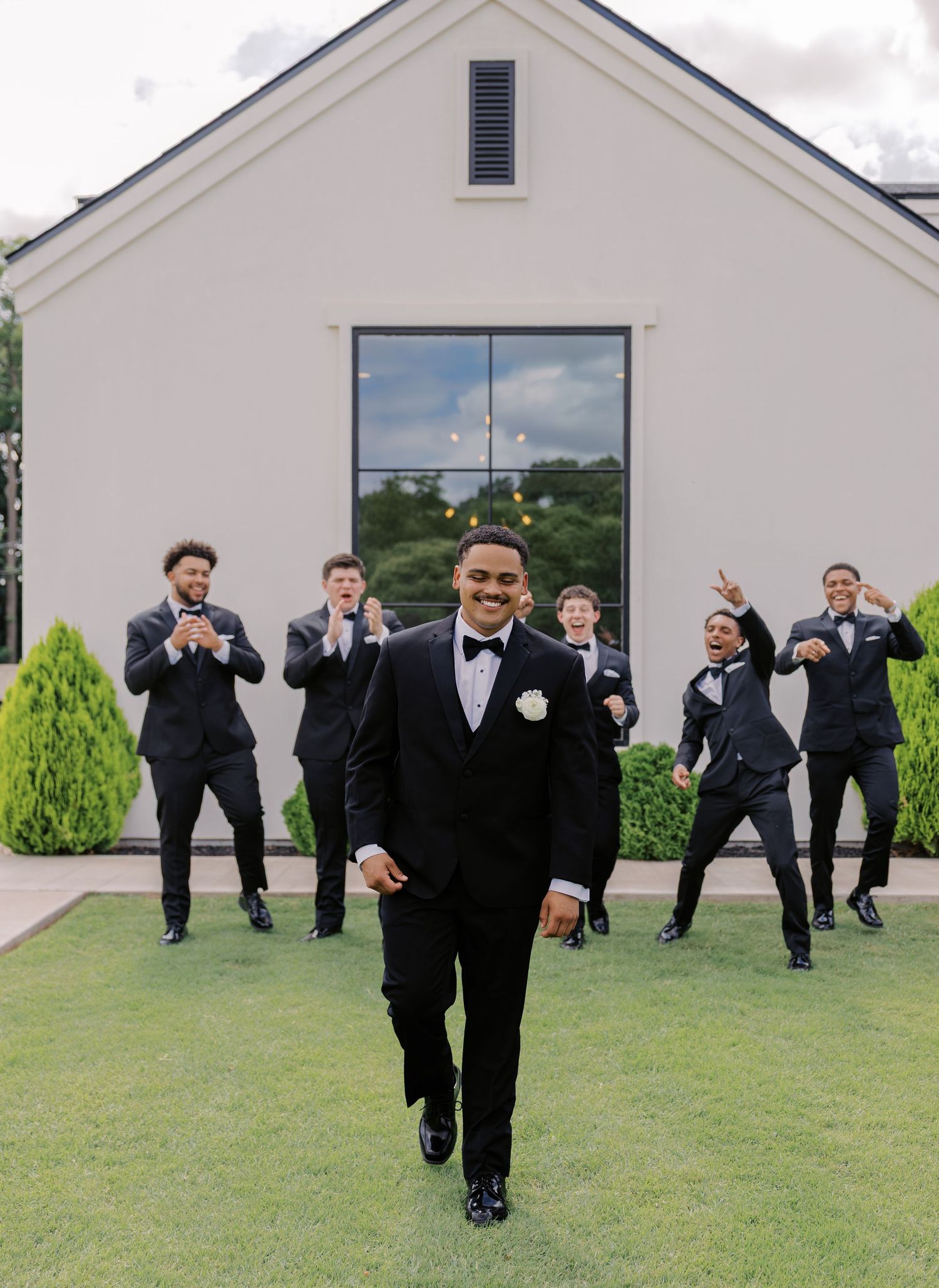 Groom in black tuxedo walks confidently toward camera while groomsmen celebrate behind him outdoors. Oklahoma wedding photographer.