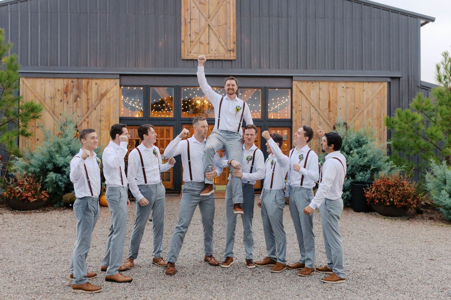 Groomsmen in white shirts and suspenders celebrate by lifting the groom outside a rustic barn venue.