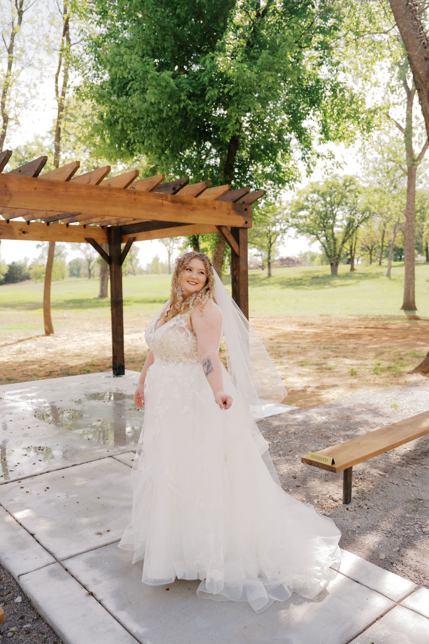 A bride in a flowing white tulle wedding gown and veil poses under a wooden pergola surrounded by lush green trees.