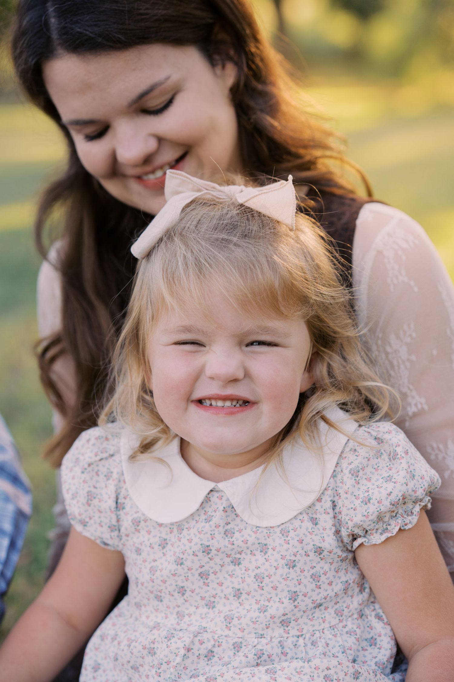 A smiling toddler girl in a floral dress with a pink bow sits with a brunette woman outdoors in a sunny park.
