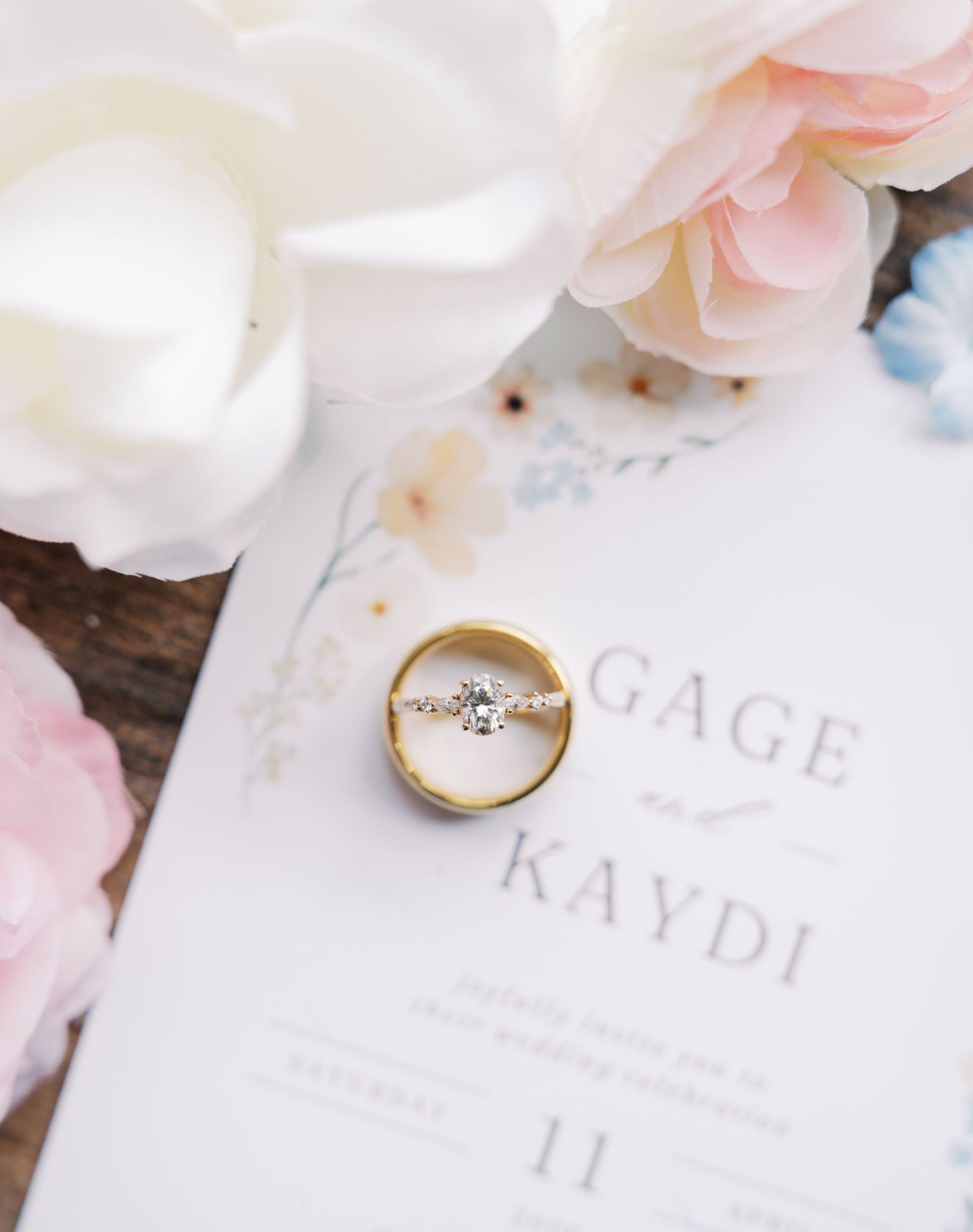 Gold wedding band and diamond engagement ring resting on a floral wedding invitation for Gage and Kaydi, surrounded by pink roses.