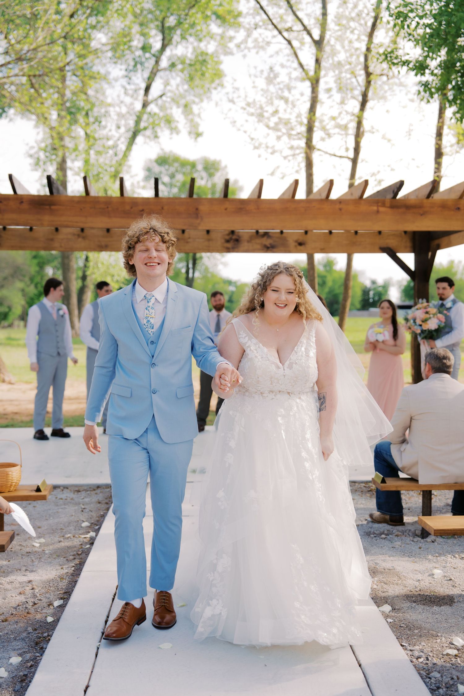 Joyful newlyweds walk down the aisle at an outdoor wedding ceremony, groom in light blue suit, bride in white gown.