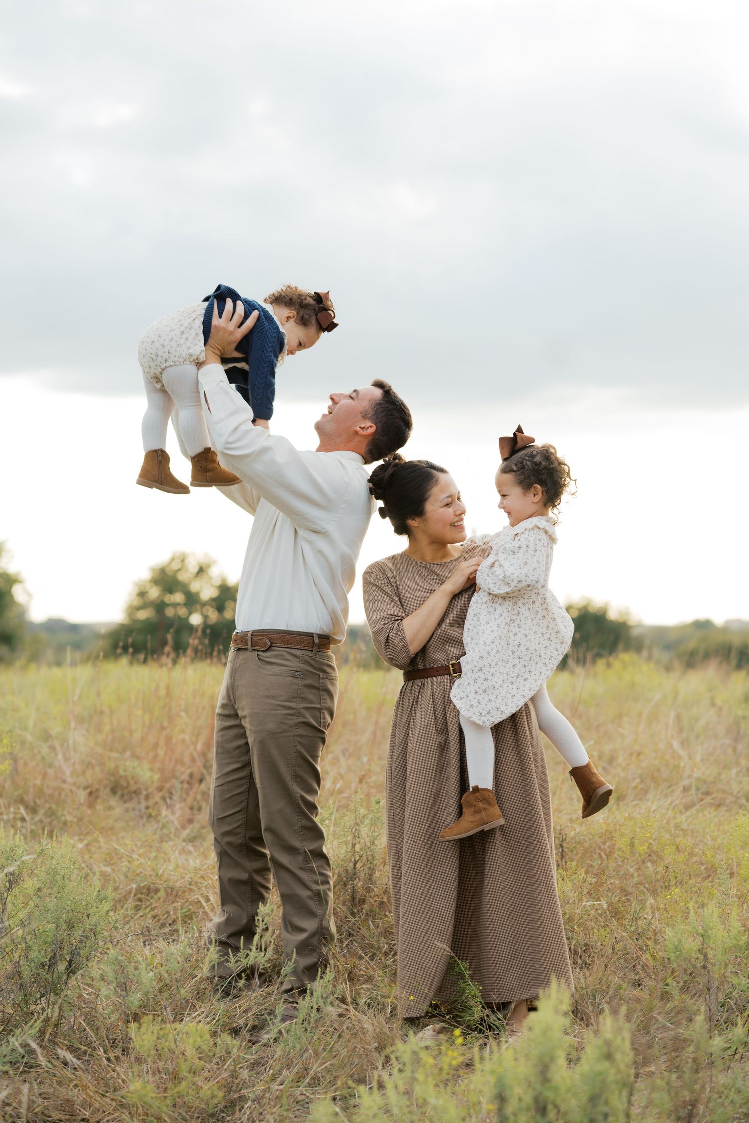 A joyful family of four plays in a field, with dad lifting a toddler and mom holding another child outdoors.
