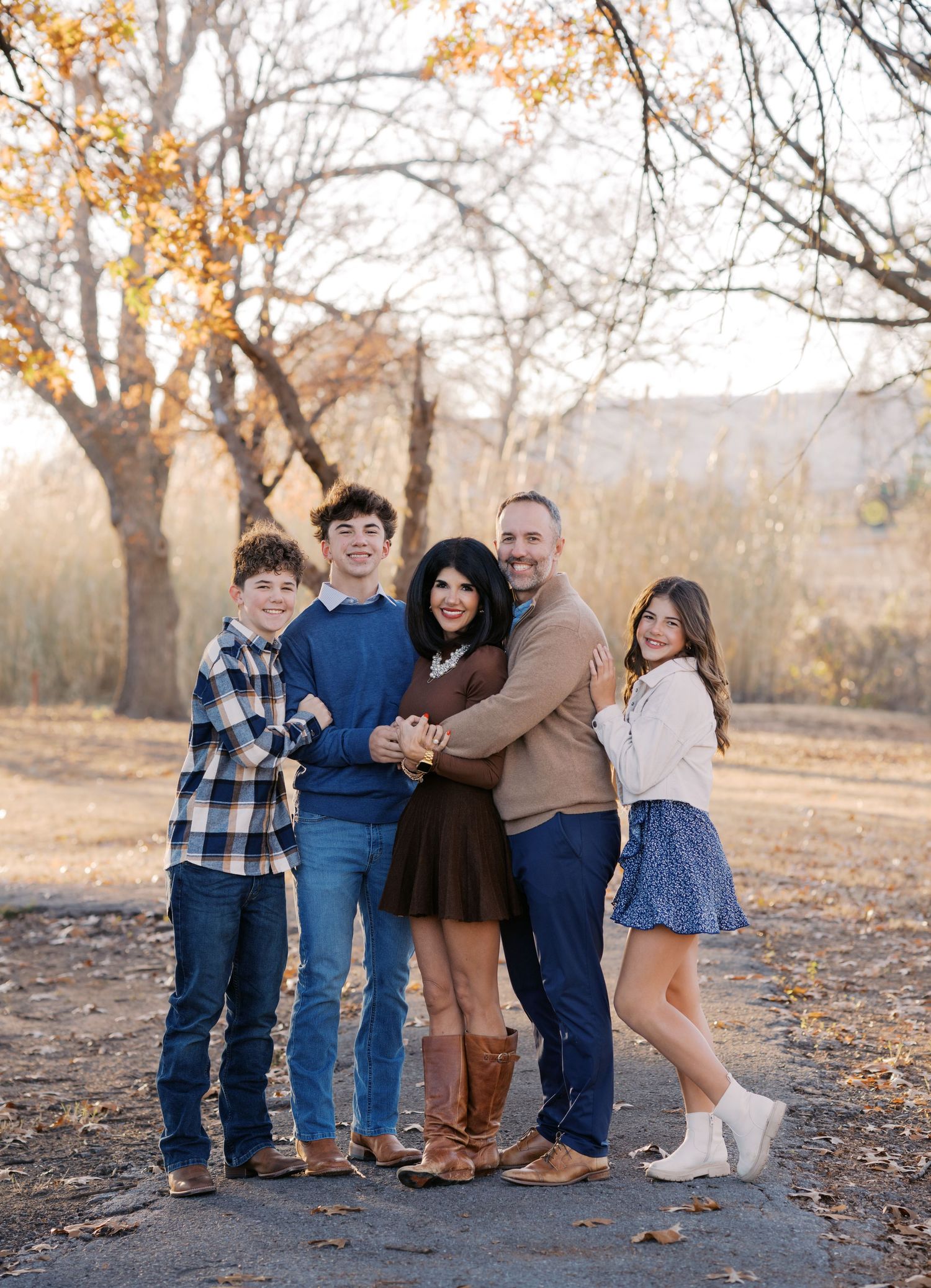 Family of five posing together outdoors in fall, wearing coordinated blue and brown outfits among autumn trees.
