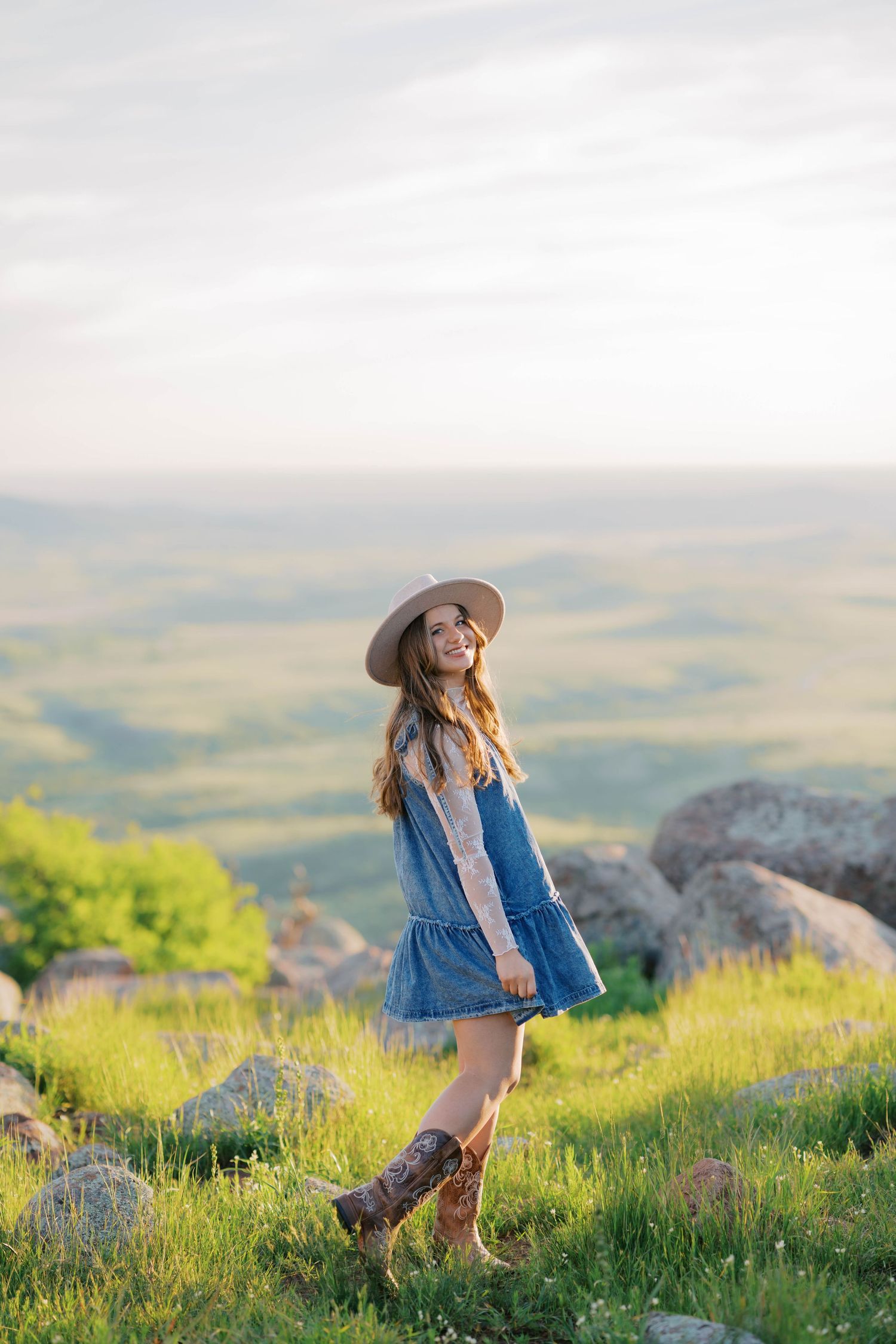 Young woman in denim dress, wide-brim hat, and cowboy boots standing on a green hillside with scenic valley views.