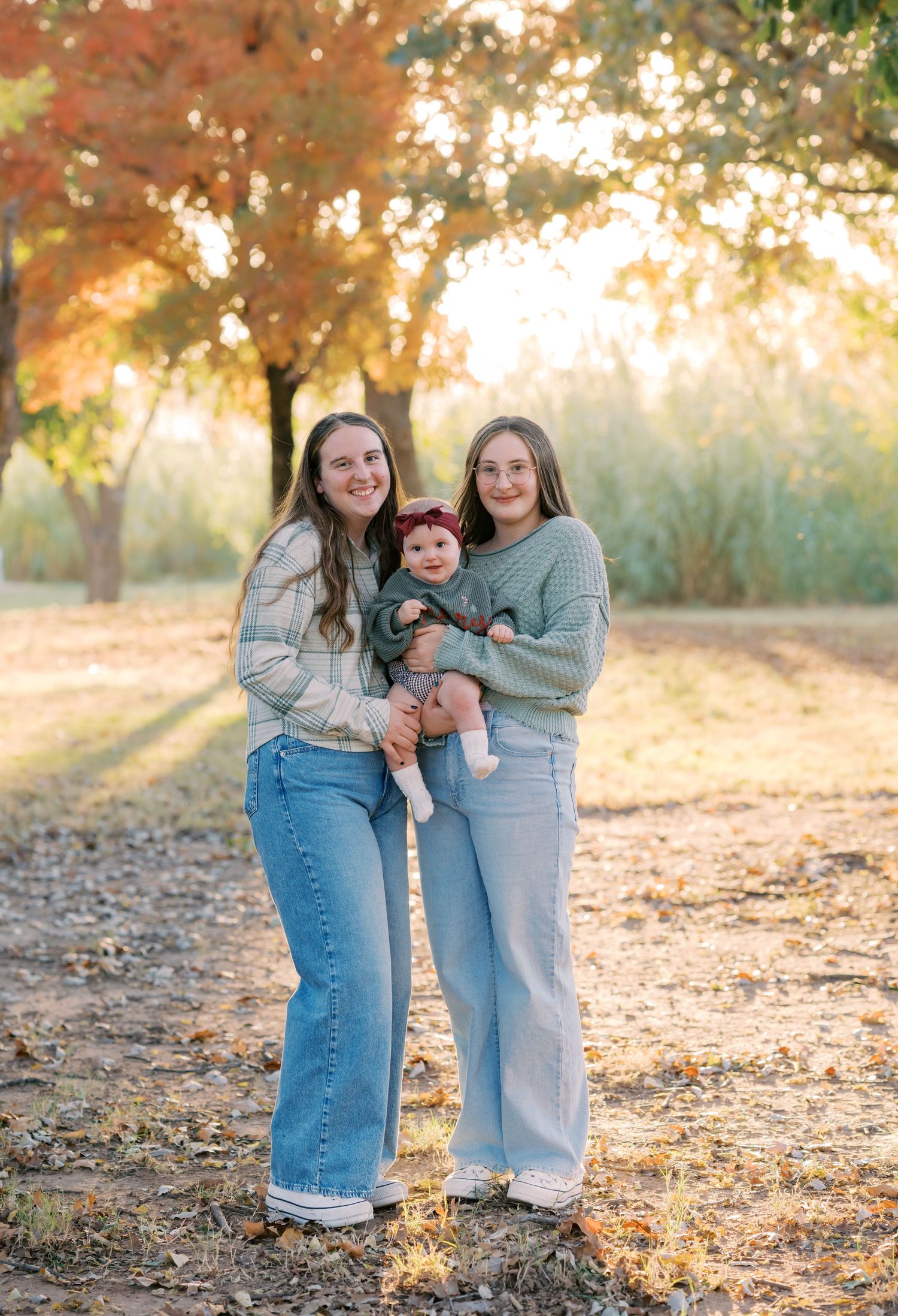 Two young women holding a baby in green outfit pose outdoors among fall foliage trees.