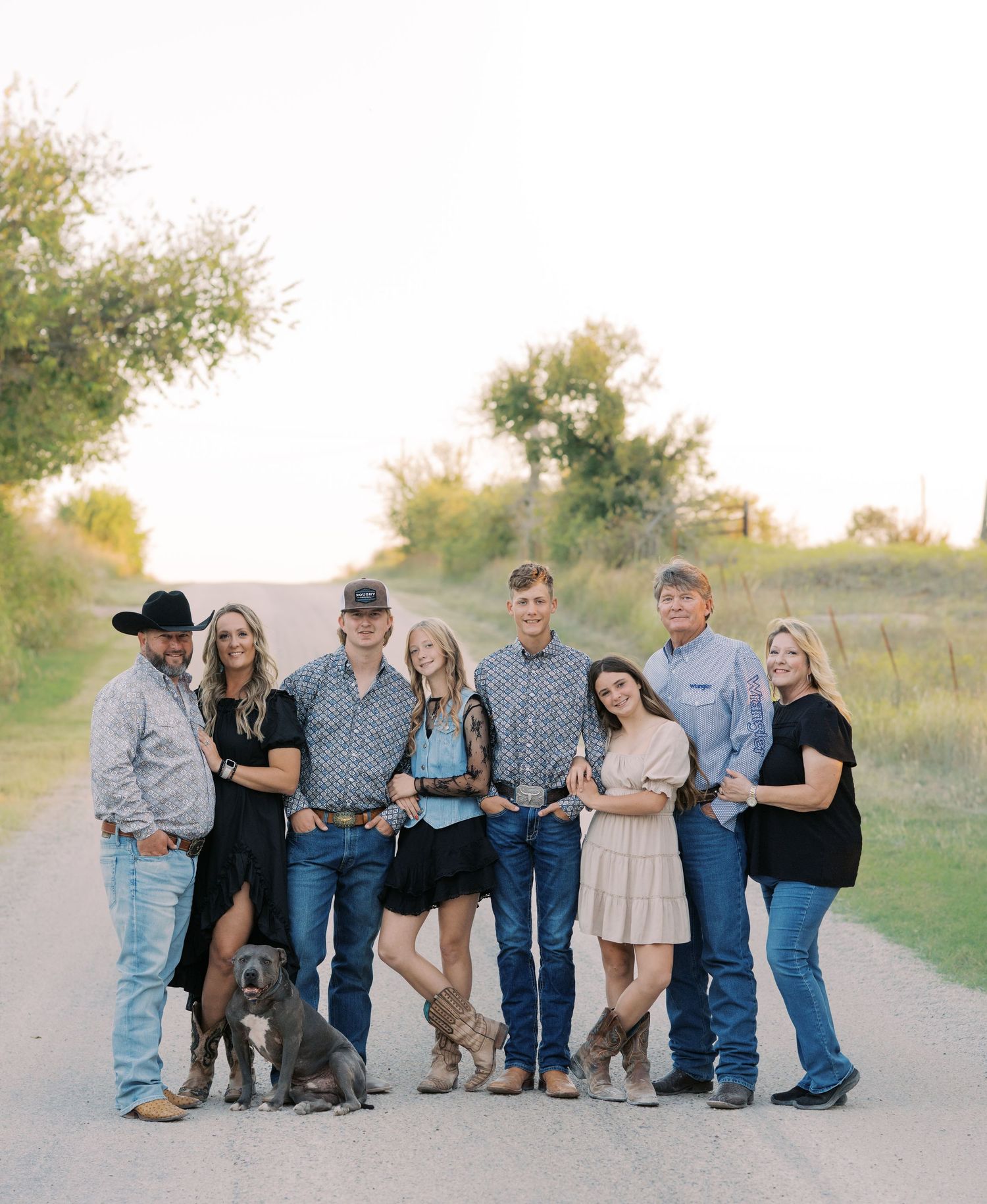 A family of eight poses with their dog on a rural dirt road at golden hour in western attire.