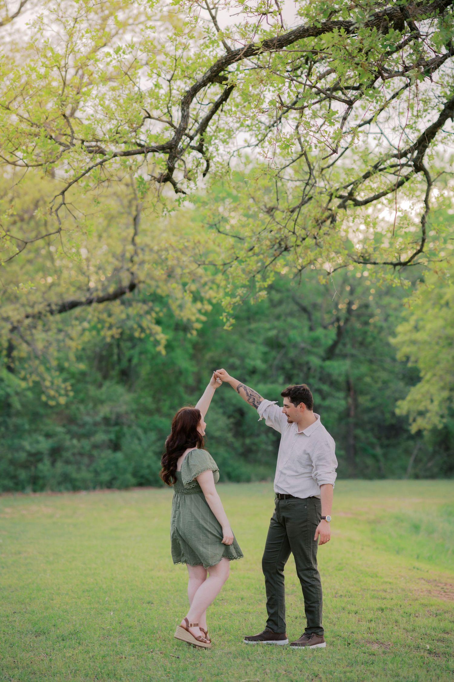 A couple dances together in a lush green park, the man spinning the woman in a green dress under a large oak tree.